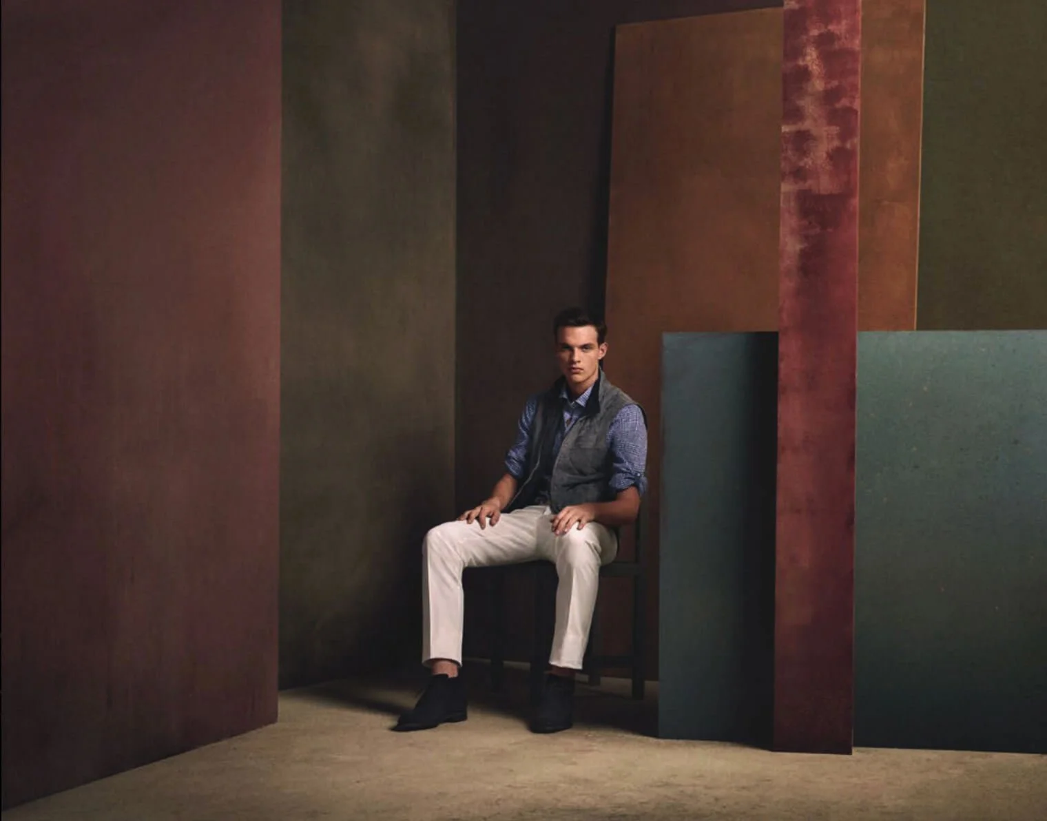 A young man sitting on a black chair against a backdrop of colorful, textured panels in shades of brown, green, and red.