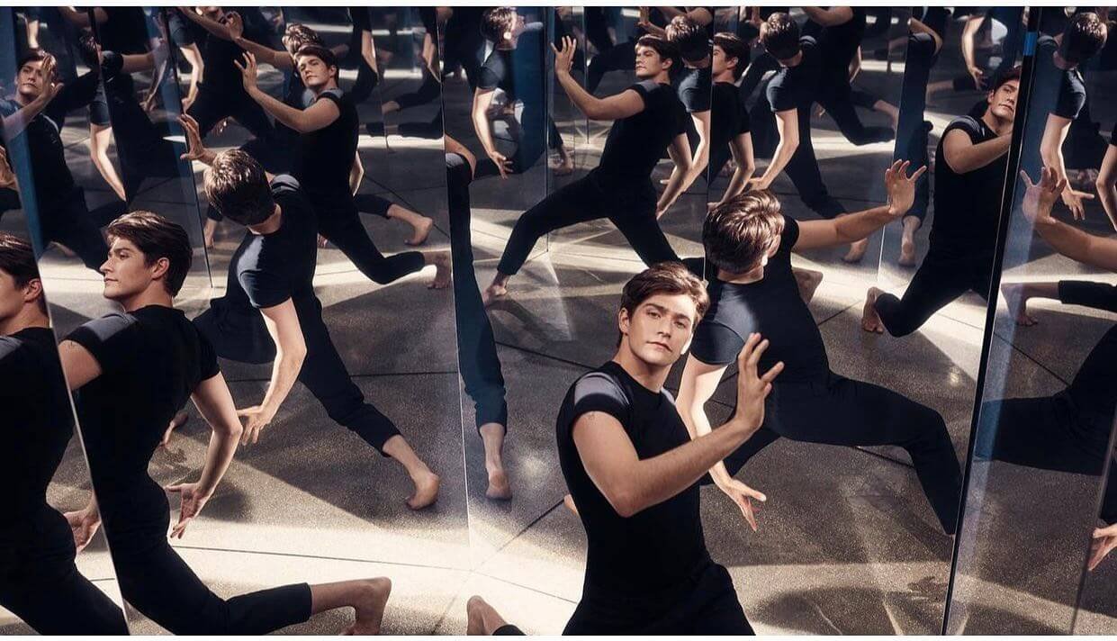 A young man in a black T-shirt and pants posing among multiple mirrors that reflect him and others in various dance or yoga poses.