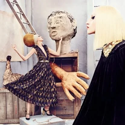 Whimsical scene featuring two mannequins and a large sculpture. The mannequin on the left is dressed in a black patterned dress with a full skirt, standing on a white platform with arms raised. The mannequin on the right has platinum blonde hair and 