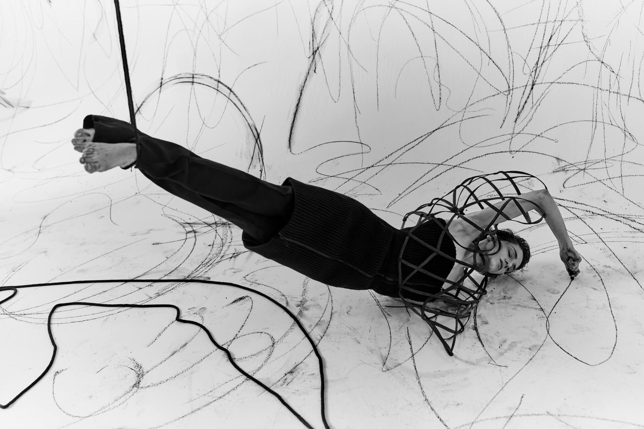 A woman lying on her side on a white floor covered with black lines and scribbles, her arm bent over her head, wearing a black dress or top, with her feet and legs exposed.