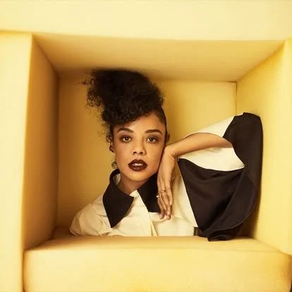 A woman with curly hair and dark lipstick poses inside a yellow box with her right arm resting on her head, wearing a beige and black outfit.
