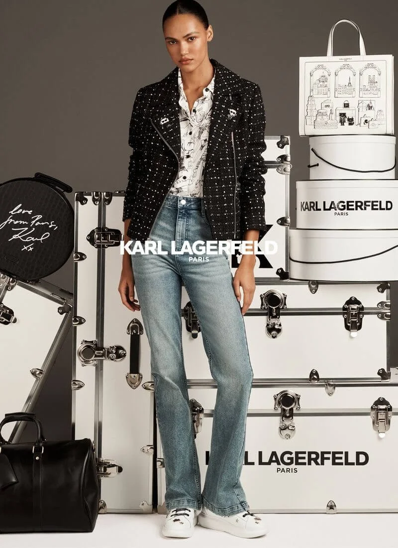 Fashion model standing in front of black and white Karl Lagerfeld display with boxes, suitcase, and bag, wearing a black tweed jacket, patterned blouse, blue jeans, and white sneakers.