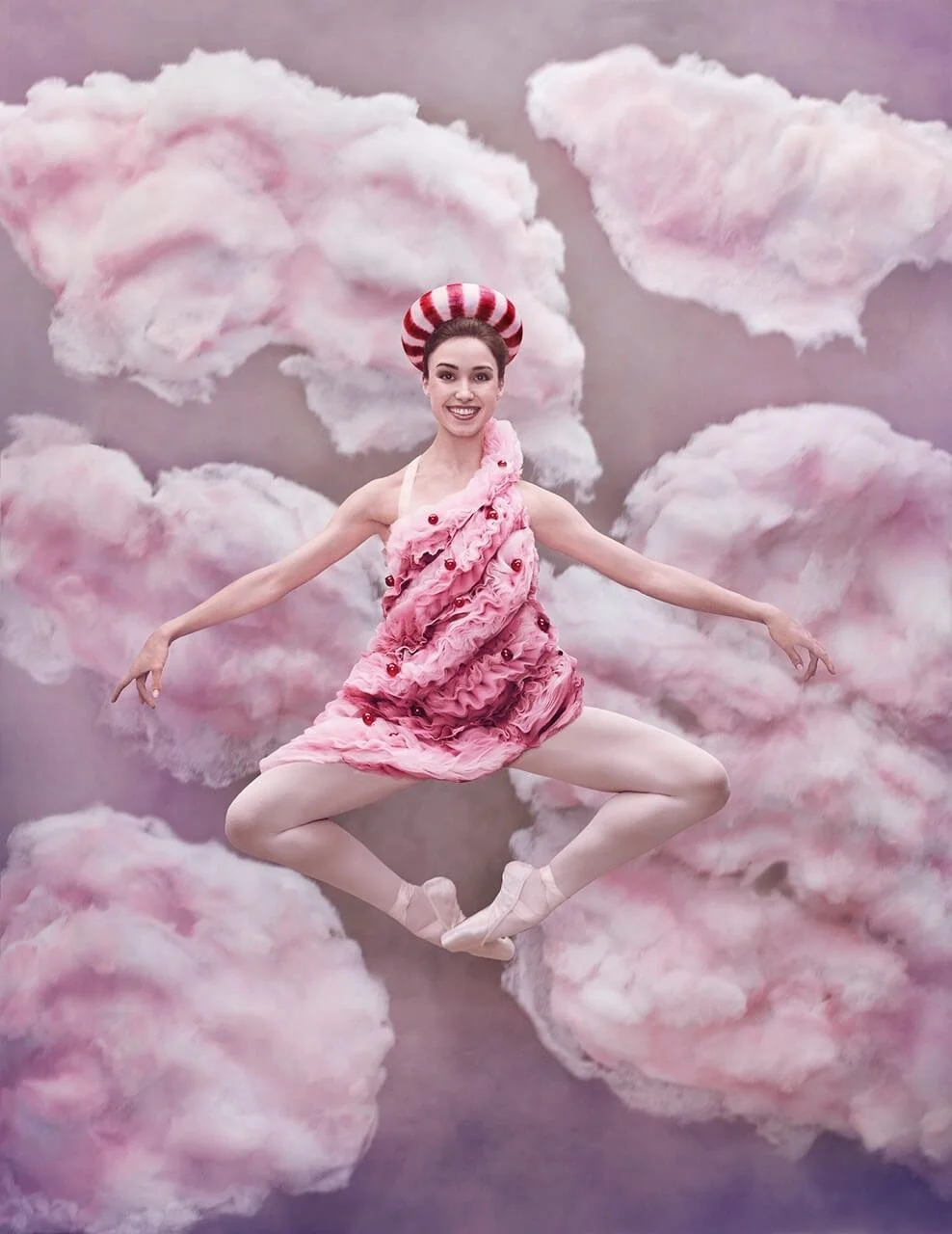 A woman in a pink dress and ballet shoes, wearing a red and white striped hat, is floating among pink and white cotton candy clouds in a surreal, whimsical setting.