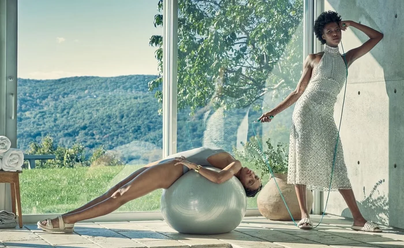 Two women in stylish dresses in a modern room with large windows showing a green outdoor landscape. One woman is lying on an exercise ball, smiling, while the other woman is standing, holding a jump rope with one hand and touching her head with the o