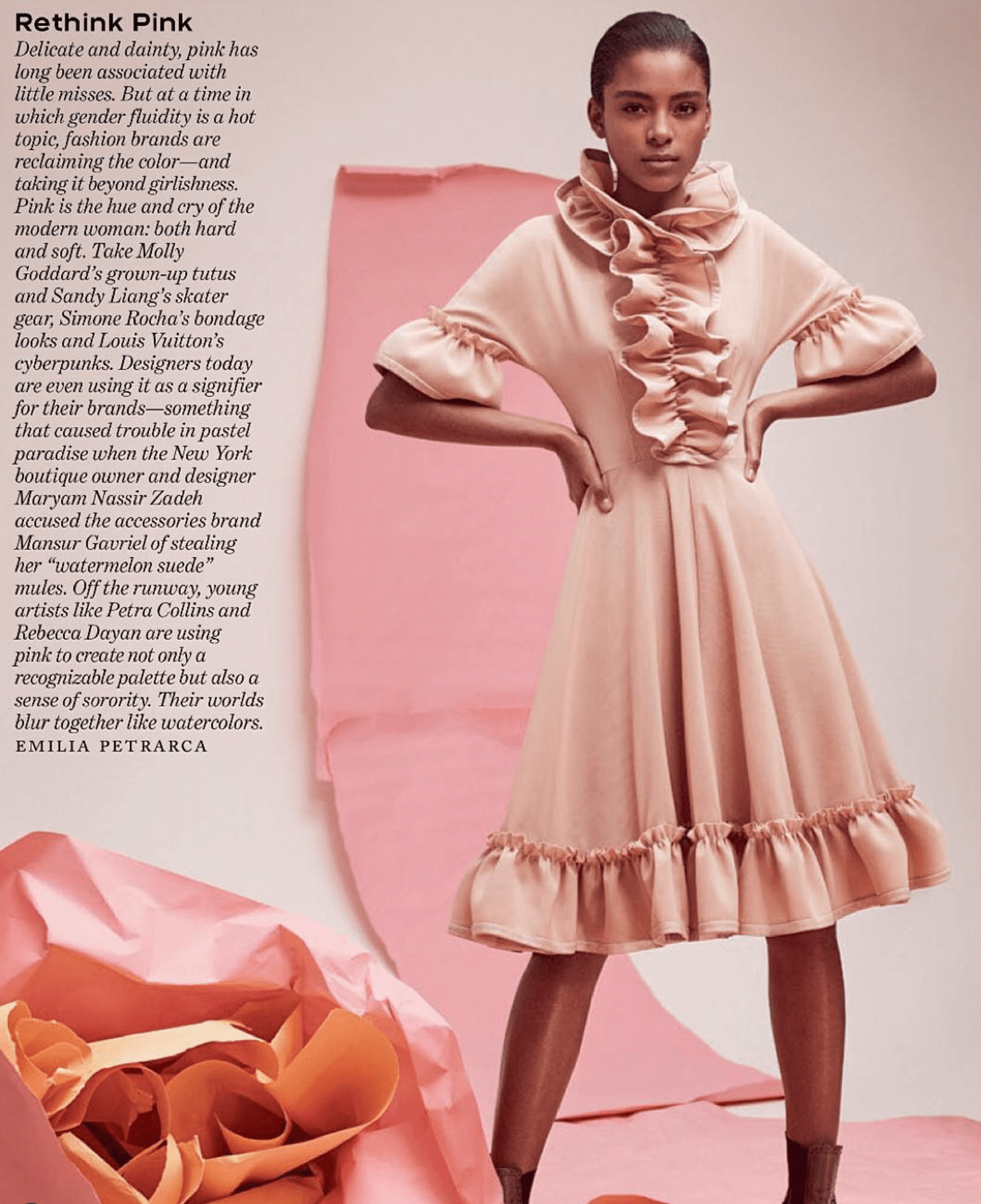 A woman wearing a pink dress with ruffles standing against a pastel pink backdrop. The dress has puffed sleeves and a ruffled collar, and the hem features a ruffled trim. To the left, there is text discussing the color pink and its cultural significa