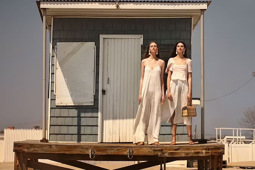 Two women standing on a small porch in front of a shed, dressed in light-colored, summery outfits, with one holding a woven handbag.