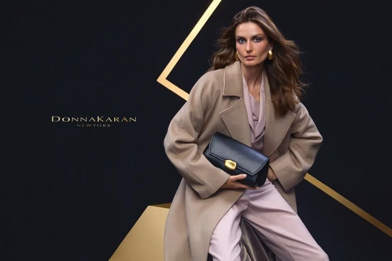 A woman in a beige coat sitting and holding a black handbag, standing against a dark background with geometric gold lines and the brand name 'DONNA KARAN NEW YORK'.