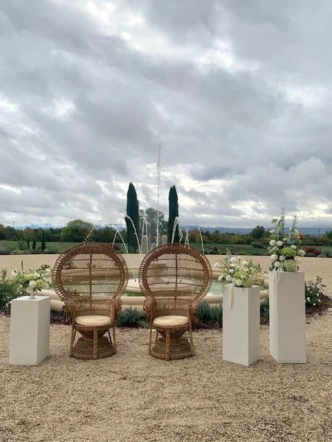 Chaises en rotin avec coussins, entourées de grands vases avec des fleurs blanches, dans un jardin avec un parterre de terre et un ciel nuageux.