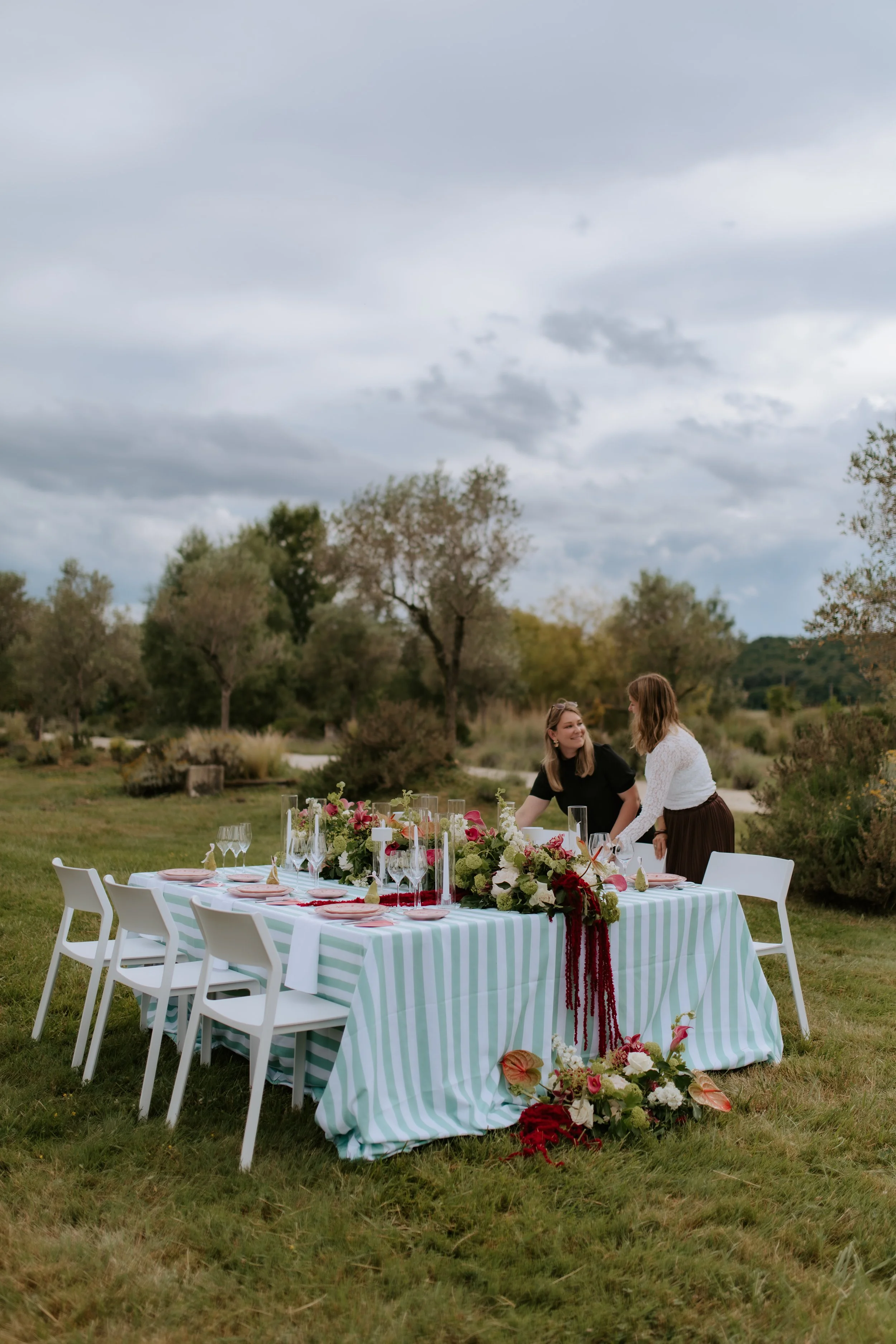 Une table décorée pour un événement en plein air, entourée de chaises blanches, avec des fleurs et des verres, dans un espace vert avec des arbres et un ciel nuageux.