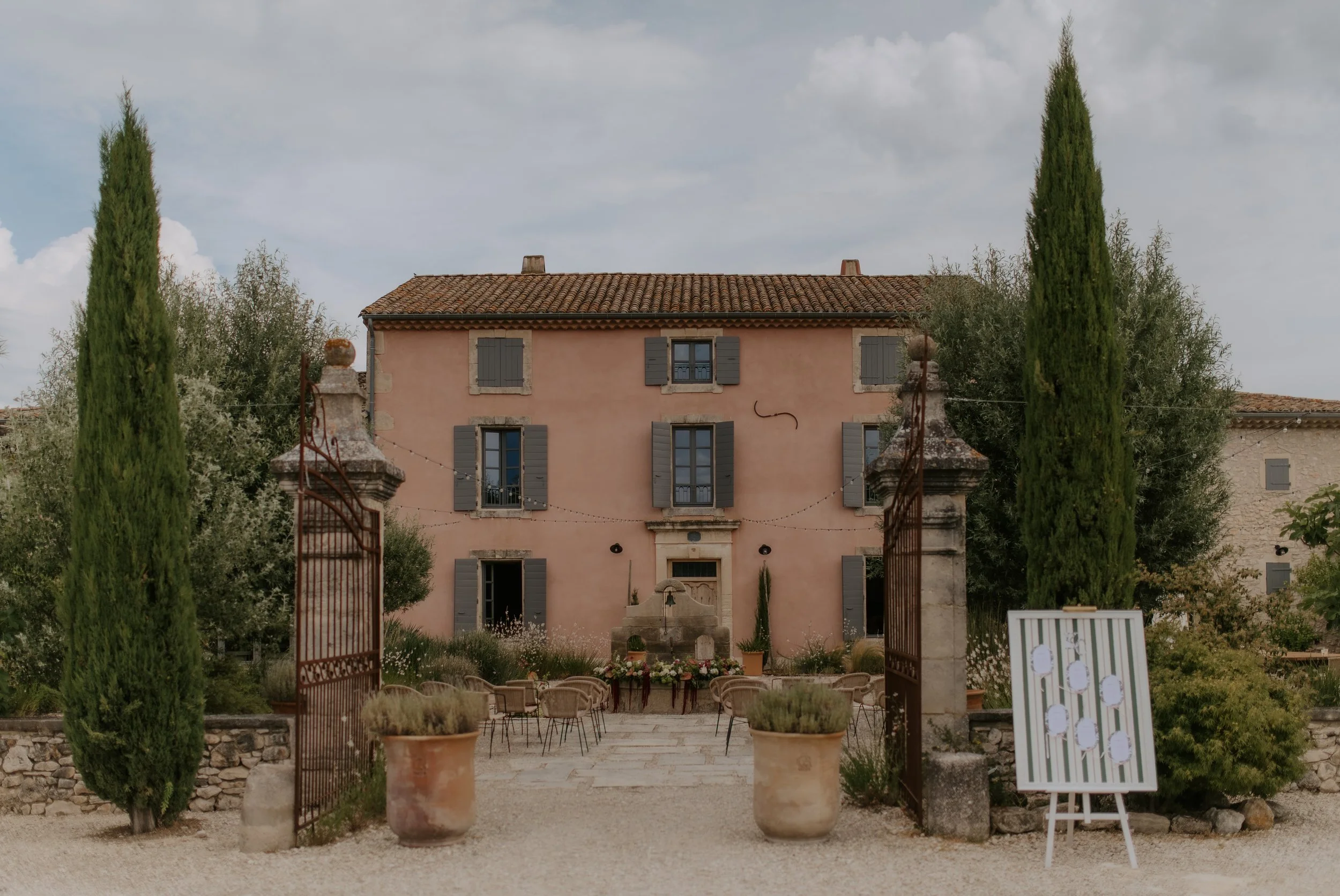 Une maison en pierre de couleur rose avec des volets en bois gris, entourée d'arbres et de plantes, avec une porte en bois et une fontaine devant. Une allée gravillonnée mène à la maison, bordée de grandes pots en terre cuite contenant des plantes.