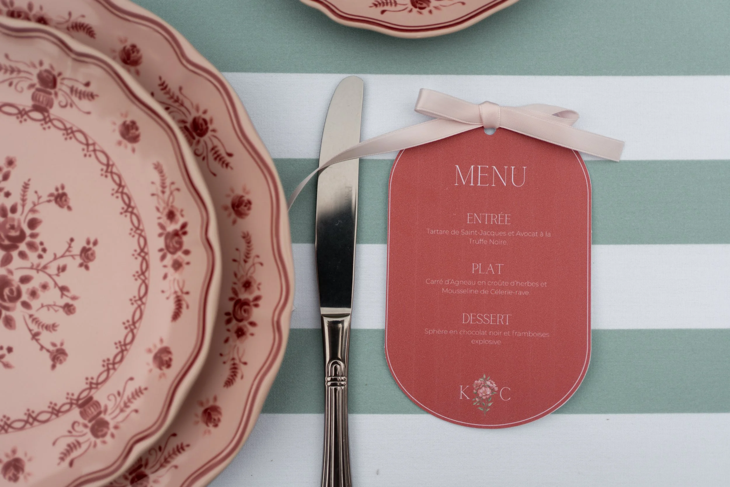 Fichier de menu de restaurant avec une assiette de vaisselle en fleurs roses, une cuillère et un couteau en métal, avec un menu en français indiquant des plats comme une tartare de Saint-Jacques, un carré d'agneau et une sphère en chocolat avec des f