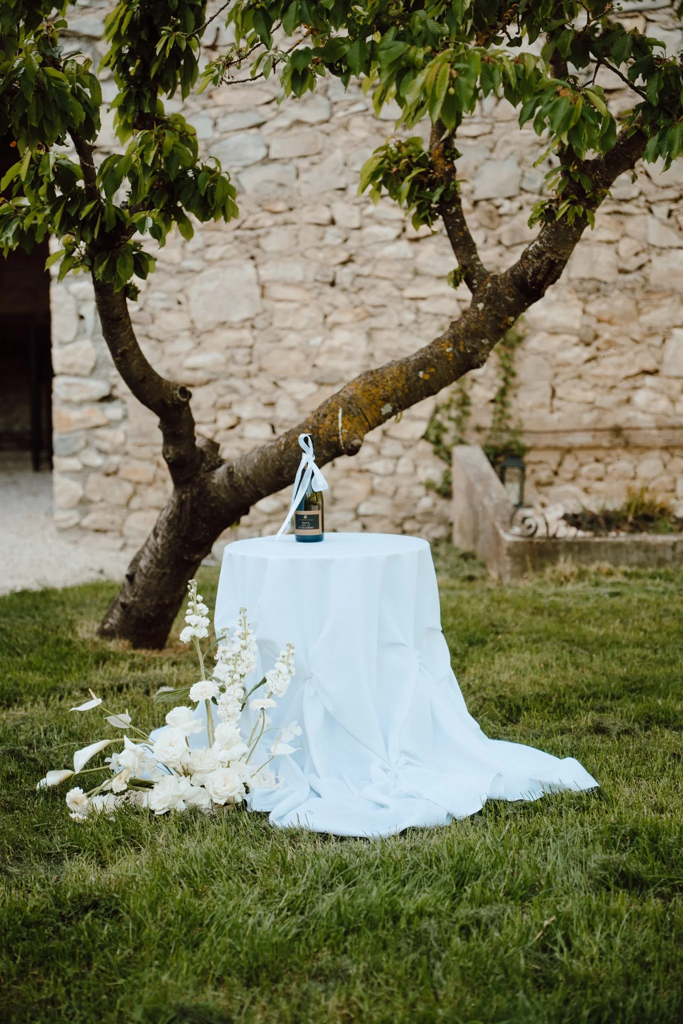Une table ronde couverte d'une nappe blanche, décorée avec un bouquet de fleurs blanches, située devant un arbre avec des feuilles vertes, et une bouteille de champagne posée sur la table, dans un jardin avec un mur en pierre en arrière-plan.