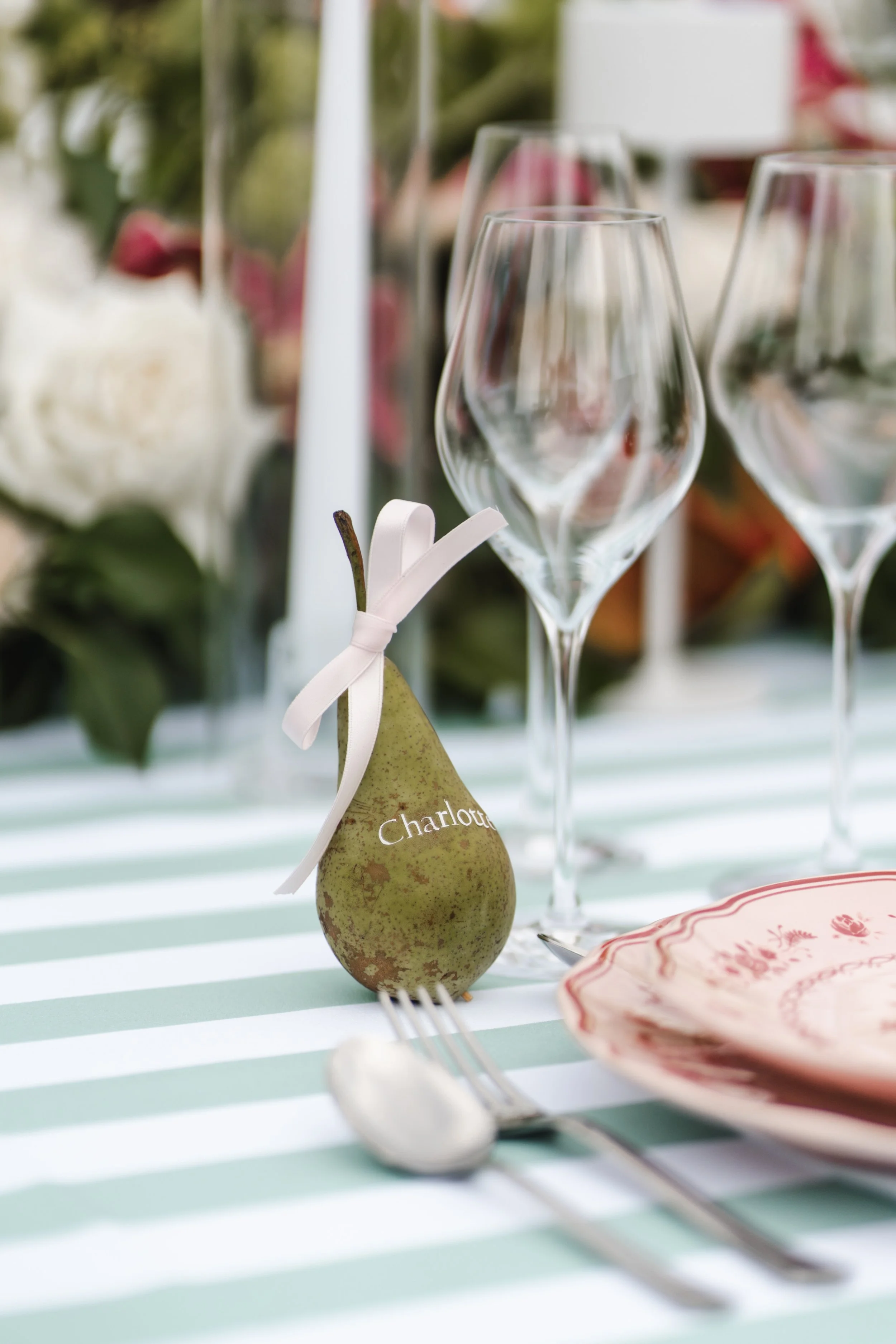 Une poire avec une étiquette portant le nom 'Charlotte' sur une table décorée pour un repas, entourée de verres à vin et d'une assiette.