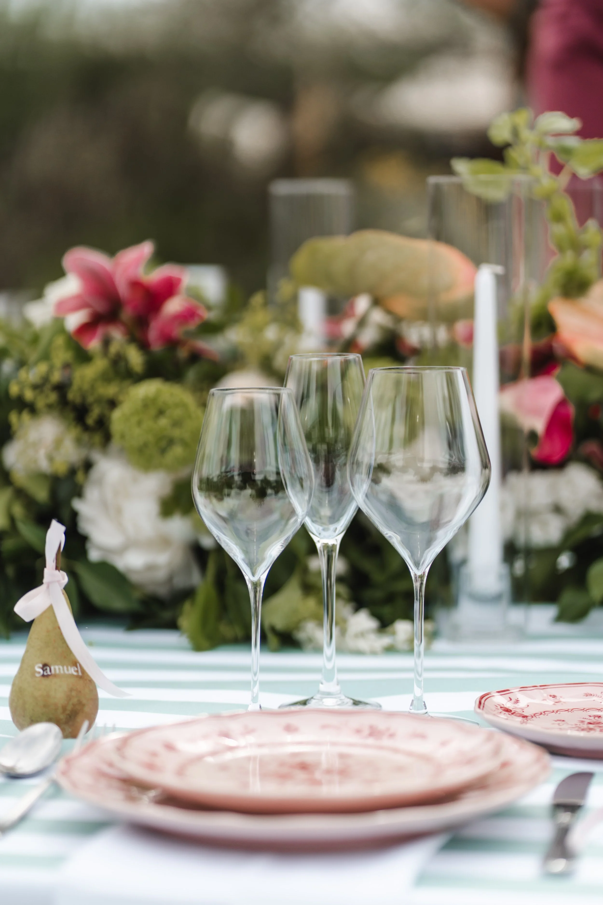 Verres à vin transparents avec une nappe à rayures vertes et blanches, décorations florales, assiettes à motifs roses et blanches, et un décor floral en arrière-plan lors d'un repas en extérieur.