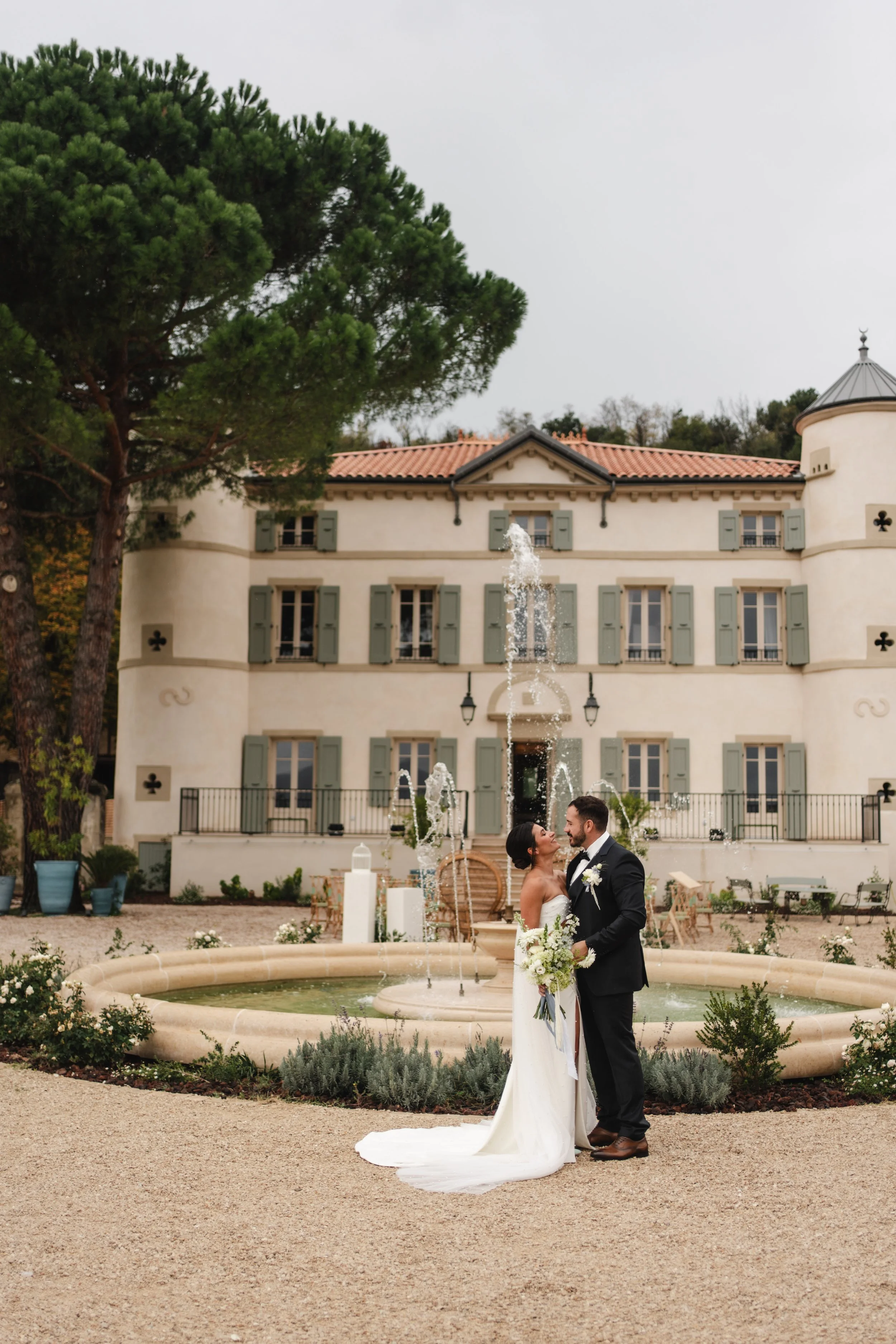 Un couple de mariés se tient devant une fontaine dans un jardin devant un grand bâtiment blanc avec volets gris et tourelles, célébrant leur mariage.