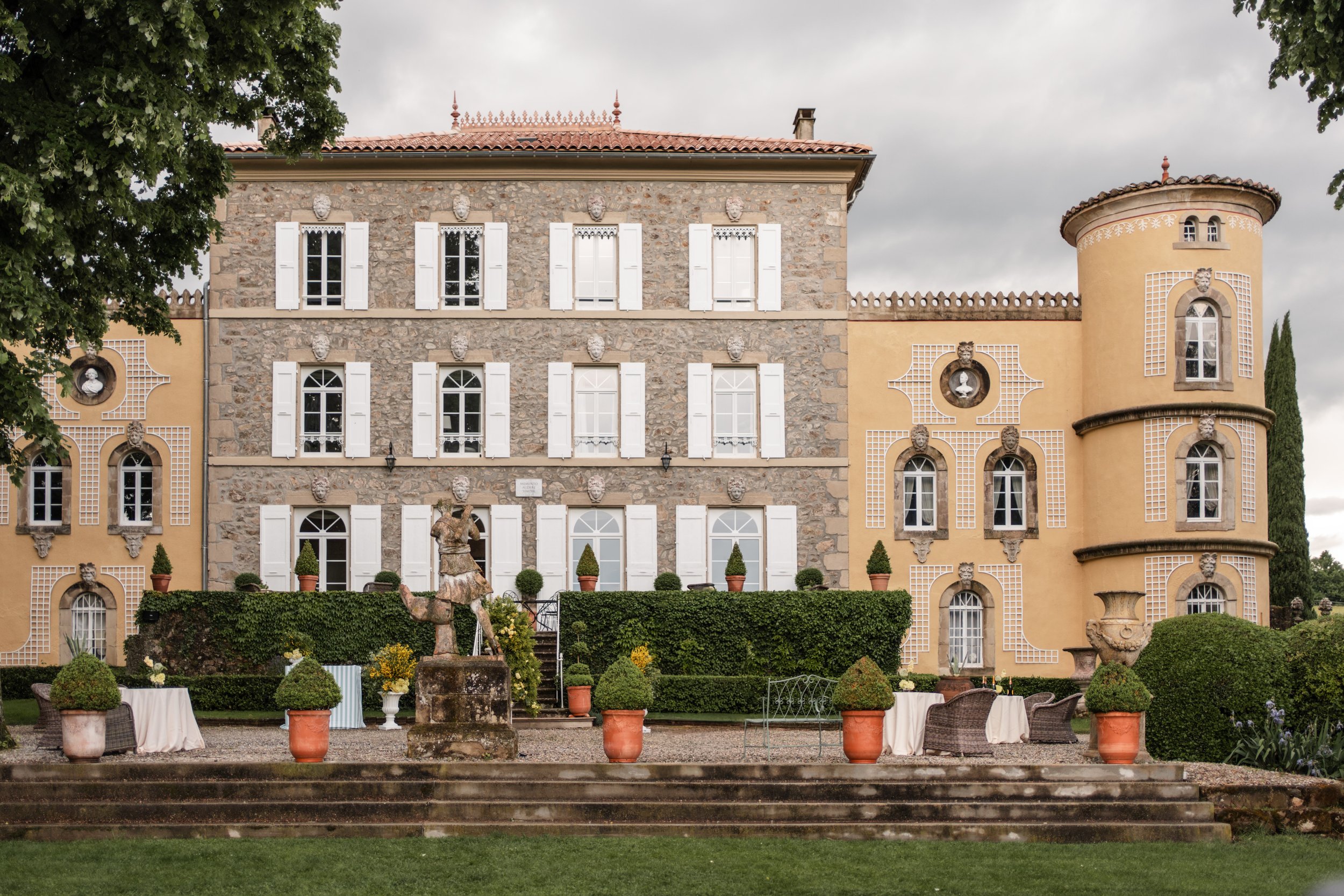 Vue de l'extérieur d'un château avec façade en pierre et murs jaunes, entouré de jardin avec statues, arbres et mobilier de jardin, ciel nuageux.