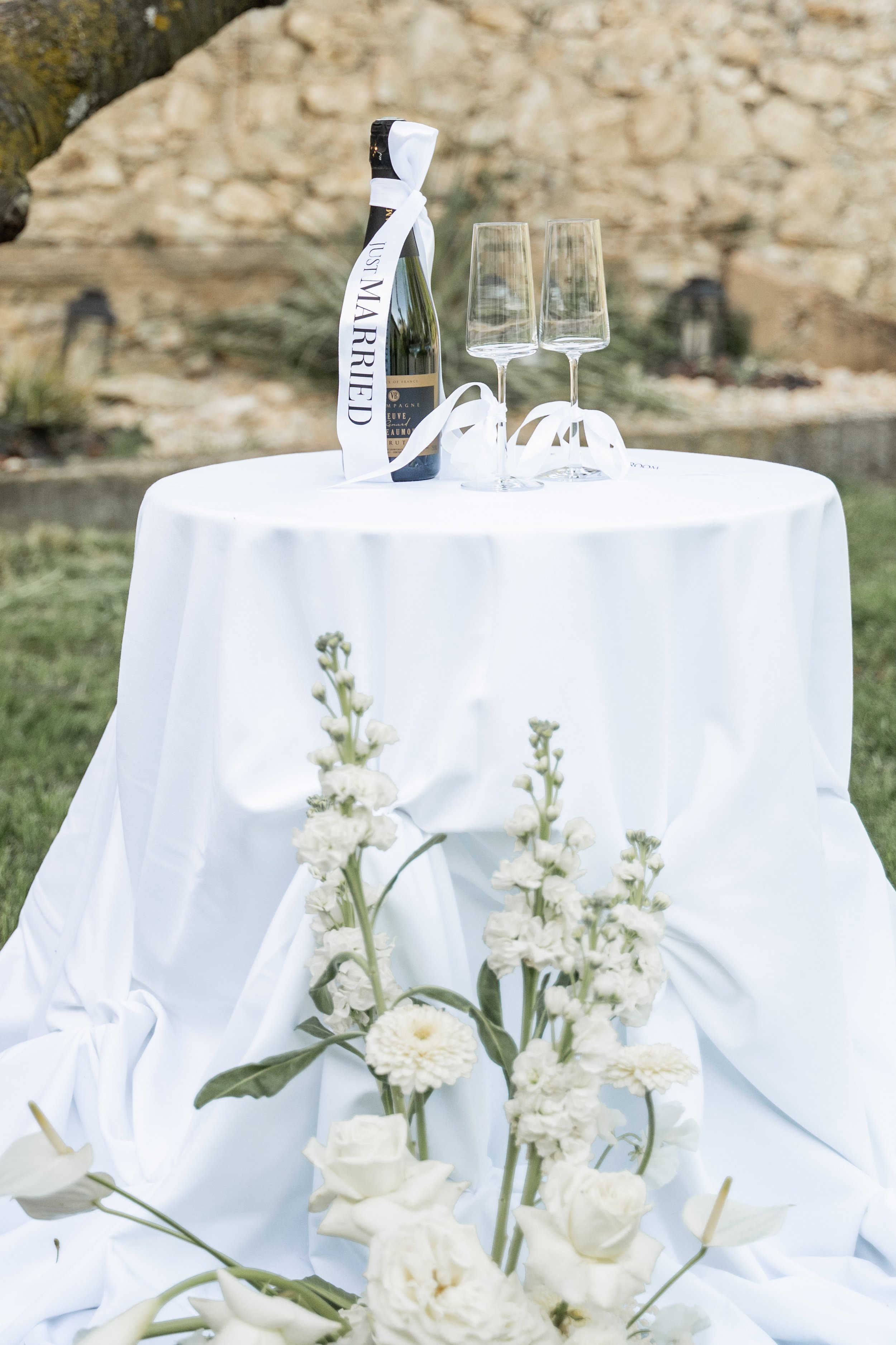 Une bouteille de champagne avec deux verres vides sur une table recouverte d'une nappe blanche, décorée de fleurs blanches, en extérieur avec un mur de pierre en arrière-plan.