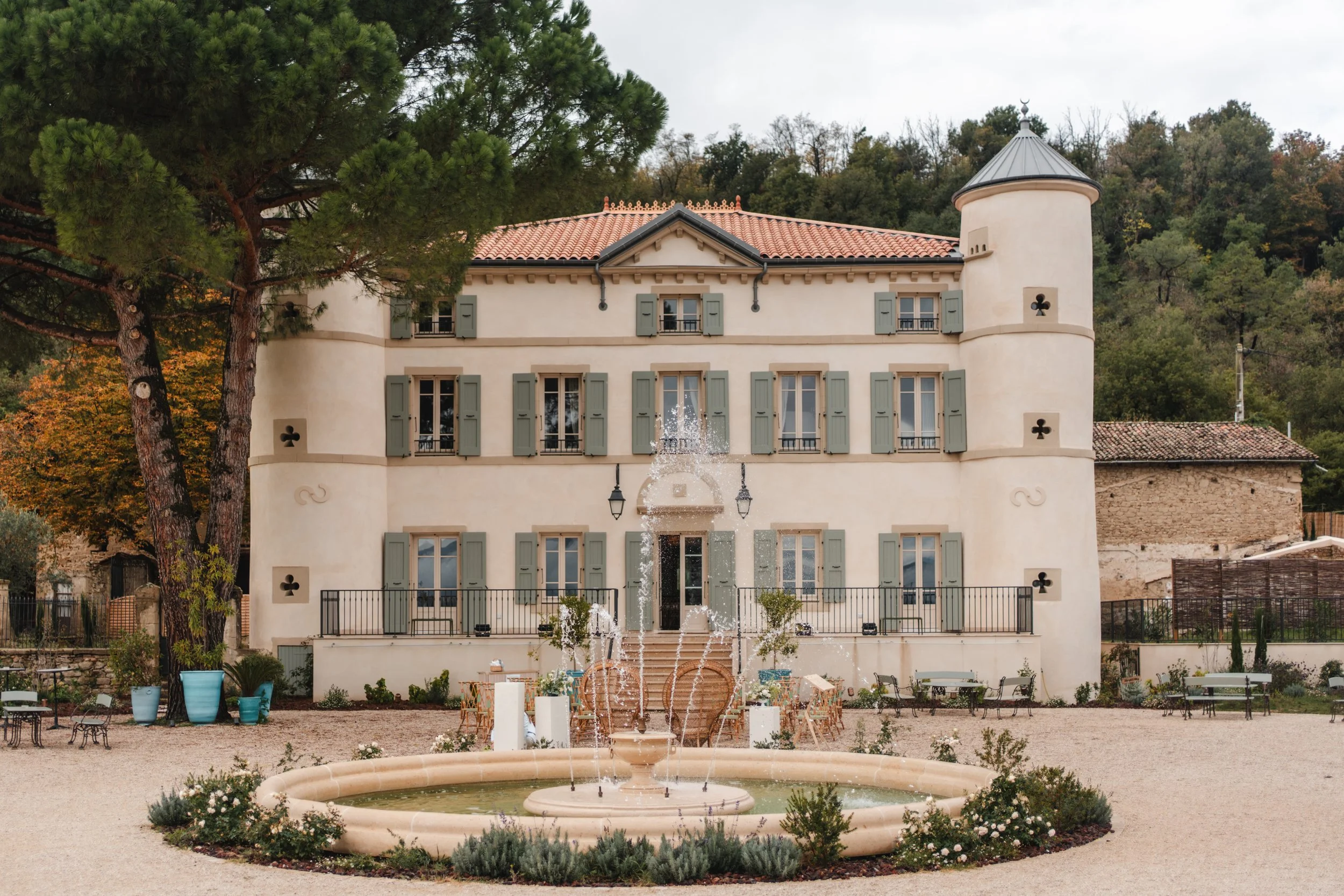 Un bâtiment de style château avec plusieurs fenêtres à volets verts, un toit en tuiles rouges et une tour ronde avec un toit conique. En premier plan, une fontaine entourée de plantes et de chaises en bois, en arrière-plan, un arbre grand avec des pi