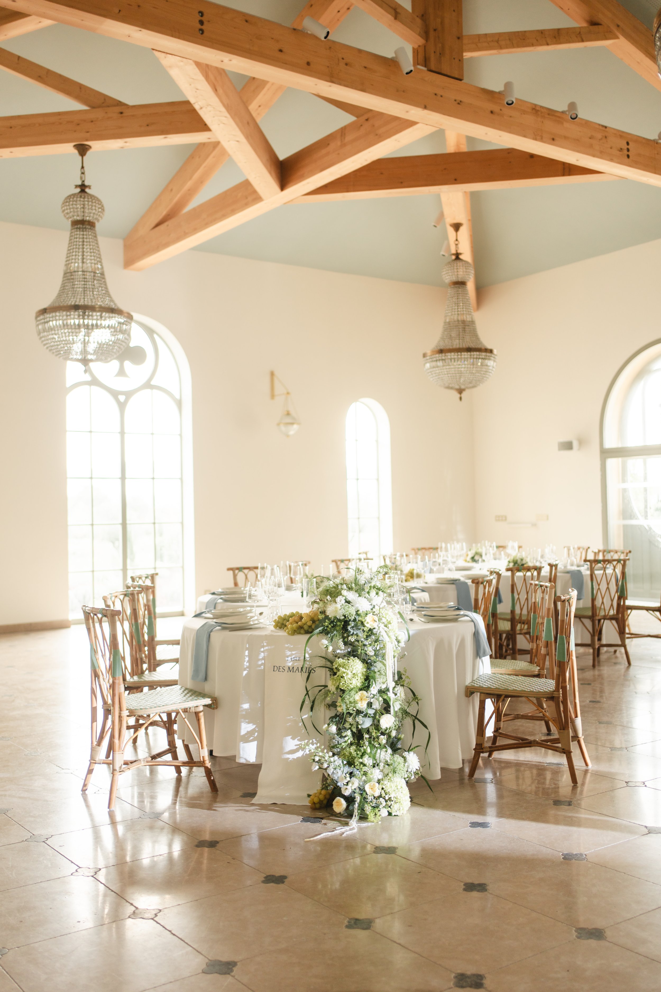 Salle de réception élégante avec tables rondes, décoration florale abondante sur une table centrale, fauteuils en bois, chandeliers en cristal suspendus au plafond avec poutres en bois apparentes, grande fenêtre en arche laissant entrer la lumière naturelle.