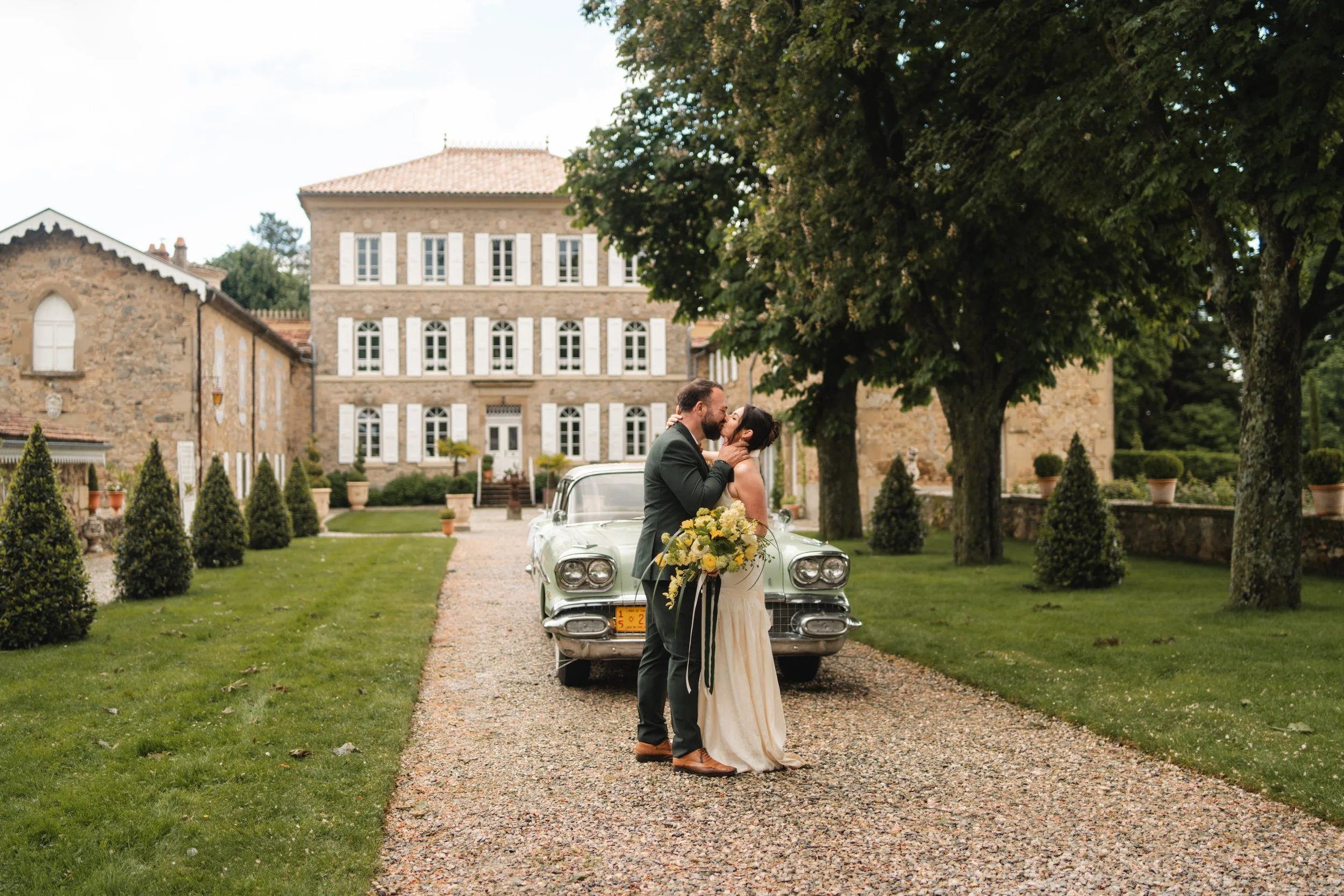 Un couple en robe de mariage s'embrasse devant une vieille voiture blanche dans une cour pavée, entourée d'arbres et de bâtiments en pierre, lors d'un mariage en plein air.