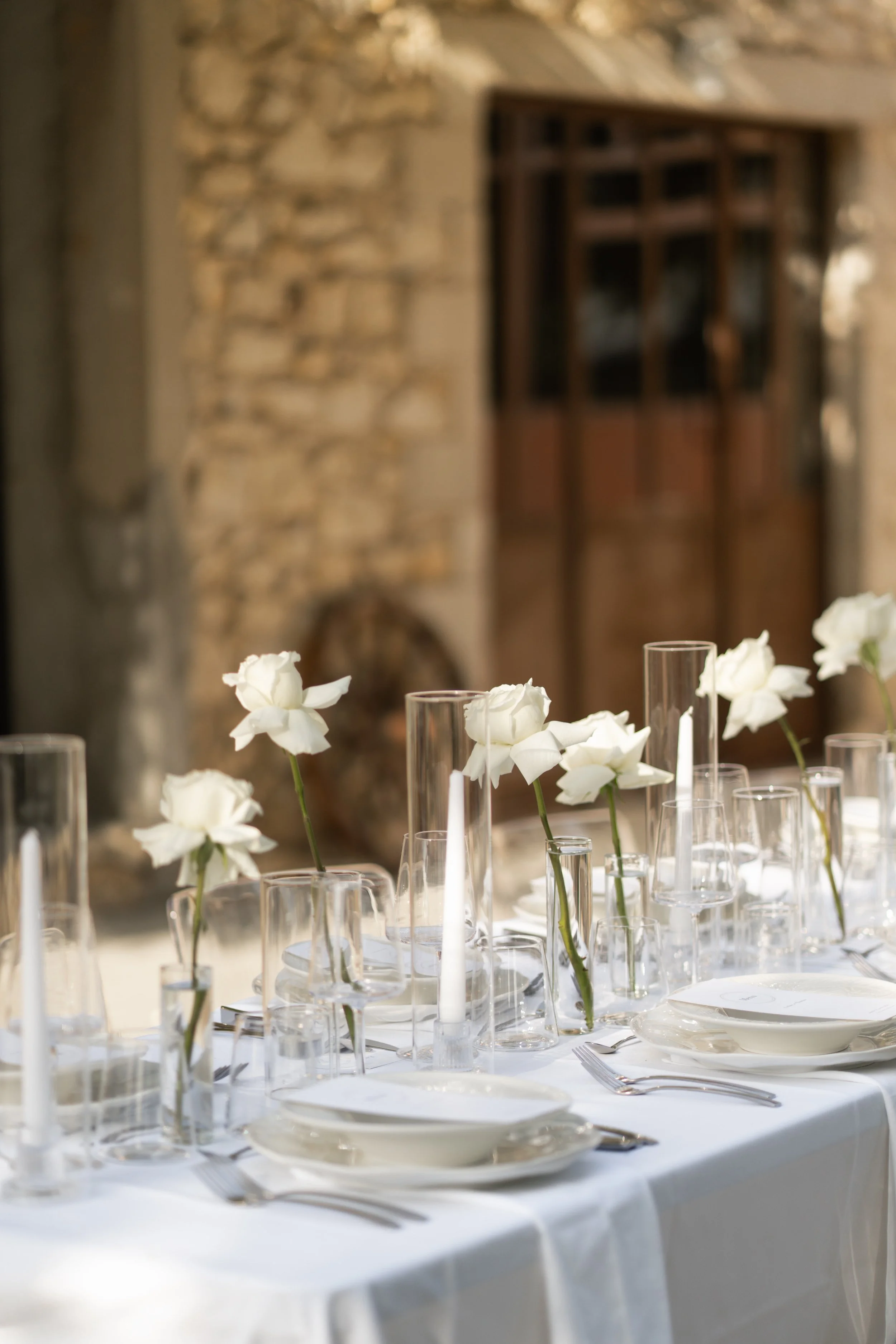 Table élégamment dressée avec des fleurs blanches dans des vases en verre, assiettes en porcelaine blanche, couverts en argent, verres à vin et bougies, décoration raffinée, ambiance chaleureuse et sophistiquée.
