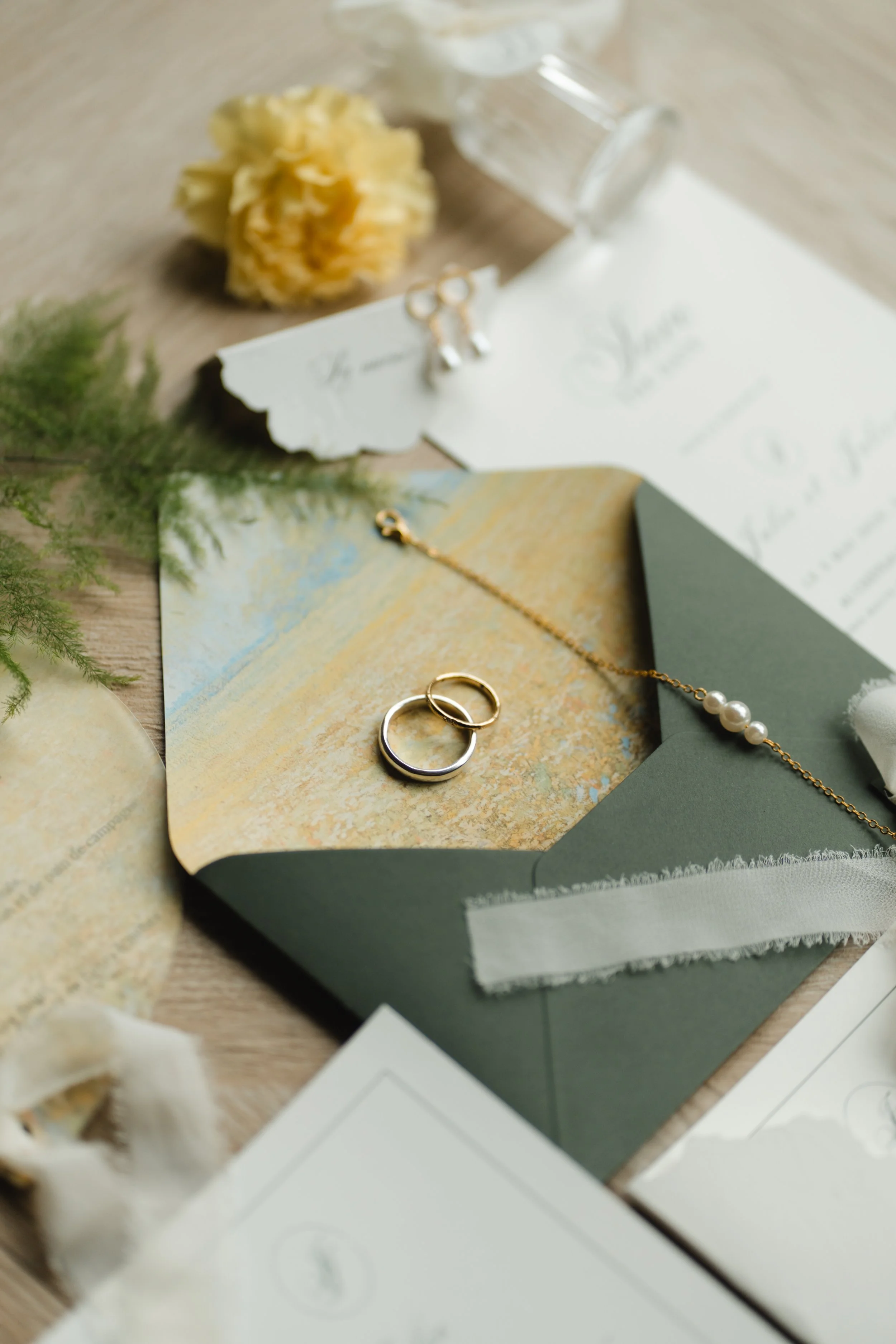 Les objets incluent des boules de fleurs, des bijoux (anneaux et collier avec perles), un verre à vin à l'envers, une carte ou une invitation, et des éléments de décoration végétale sur une table en bois.
