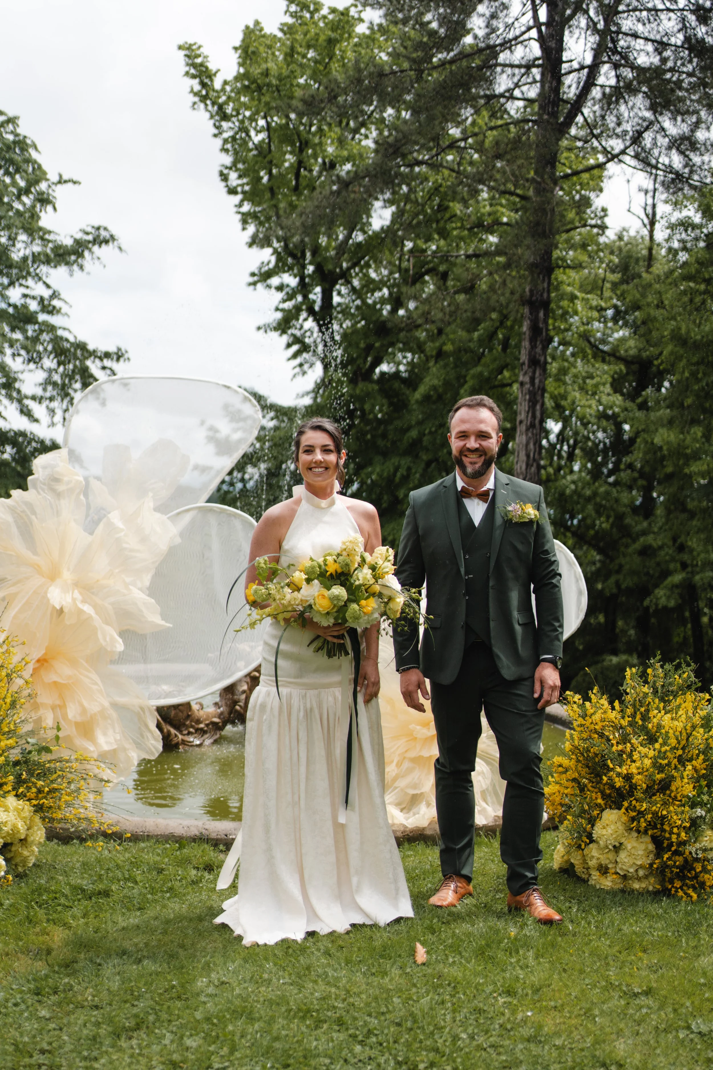 Un couple de mariés marche dans un jardin lors d'un mariage, entouré de fleurs jaunes et de structures décoratives en brut. Elle porte une robe blanche et tient un bouquet de fleurs, lui porte un costume sombre avec un nœud papillon.