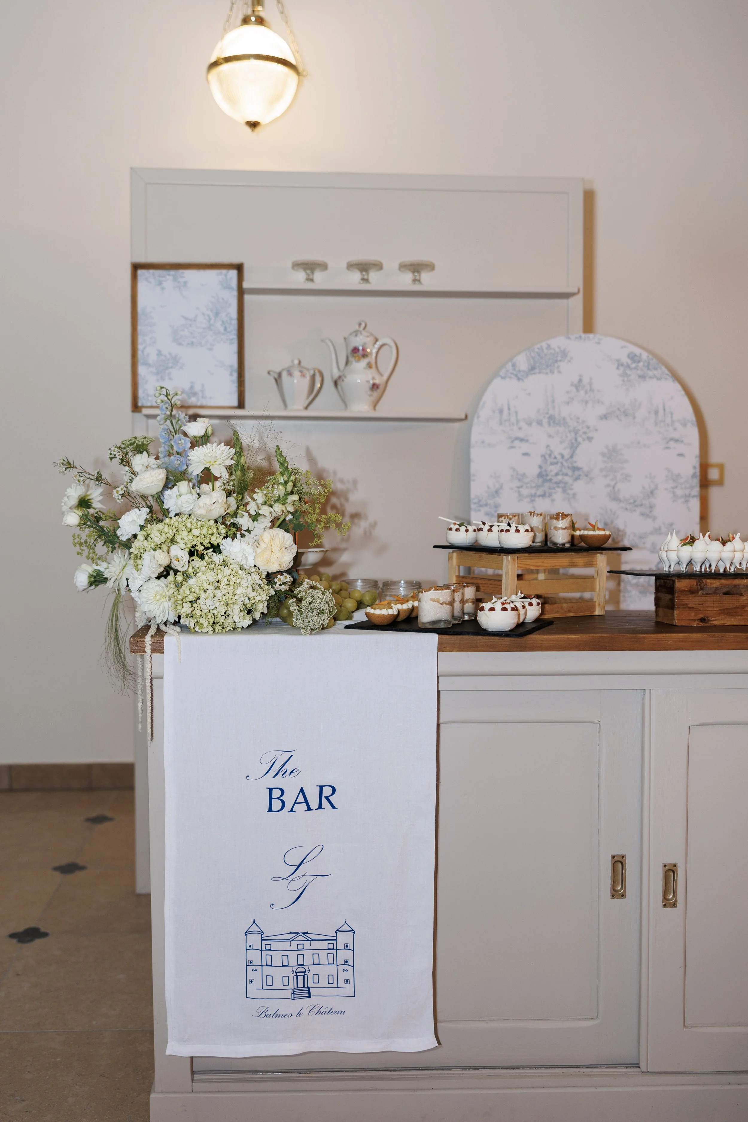 Un comptoir de bar décoré avec un grand bouquet de fleurs blanches et pastel, des desserts en verrines, et un panneau blanc avec un dessin d'un château et le texte 'The BAR' en bleu. En arrière-plan, un mur blanc avec étagères portant de la vaisselle