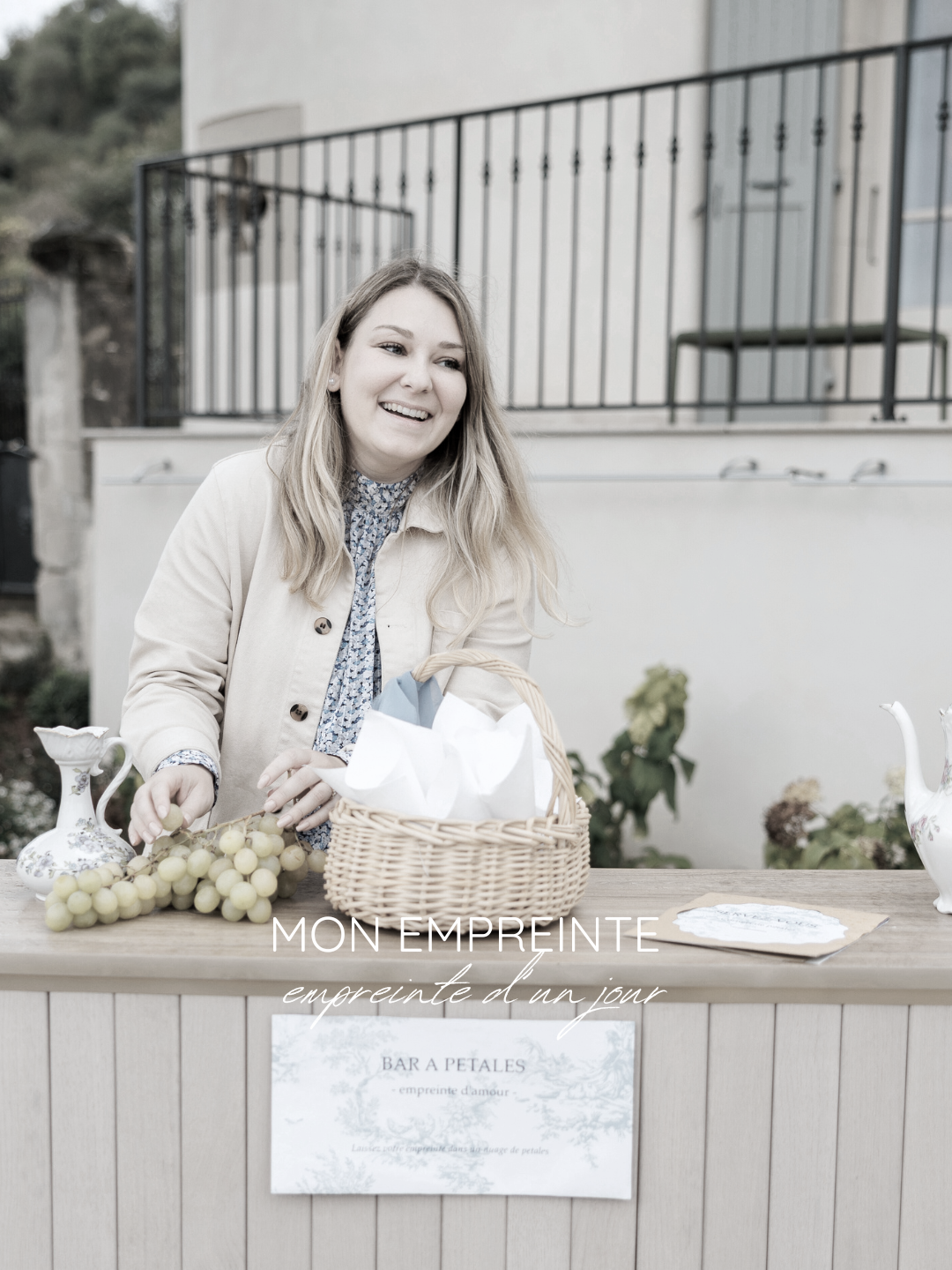 Une femme souriante derrière un stand avec des raisins verts, une carafe et un panier, lors d'un événement en plein air.