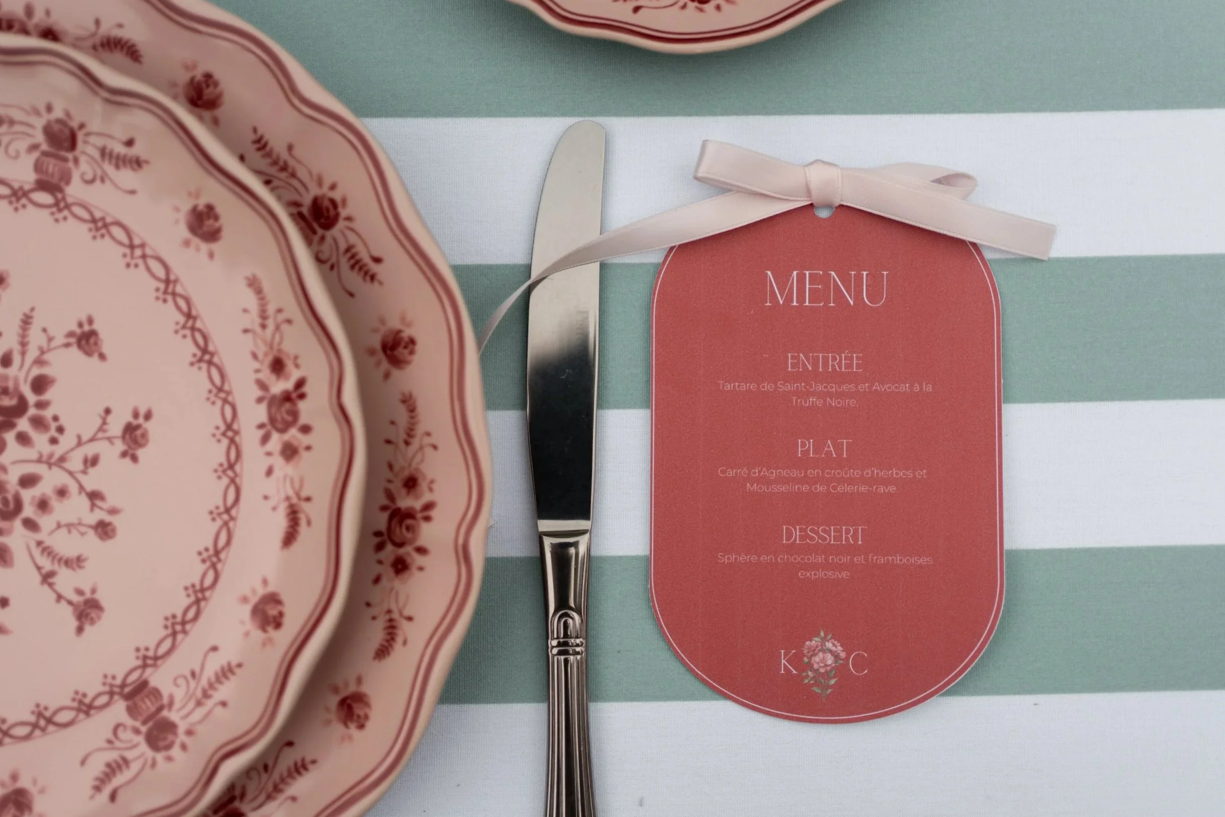 Une table avec une assiette en porcelaine rose à motif floral, un couteau en métal, et un menu avec une attache en ruban rose sur fond rayé vert et blanc.
