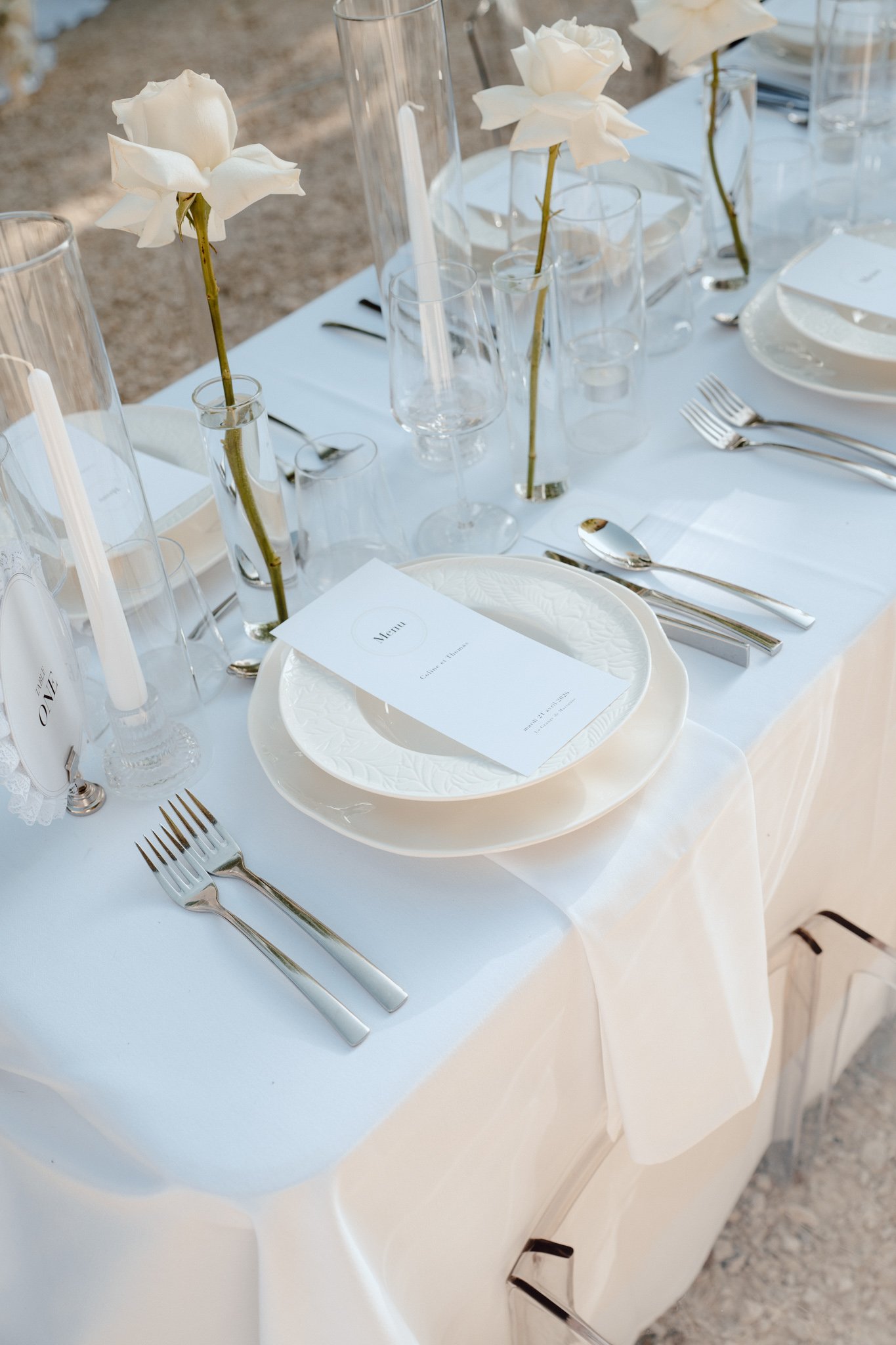 Table de mariage élégante avec vaisselle blanche, verres, couverts en acier inoxydable, fleurs blanches en vases, bougies et menu sur une assiette.