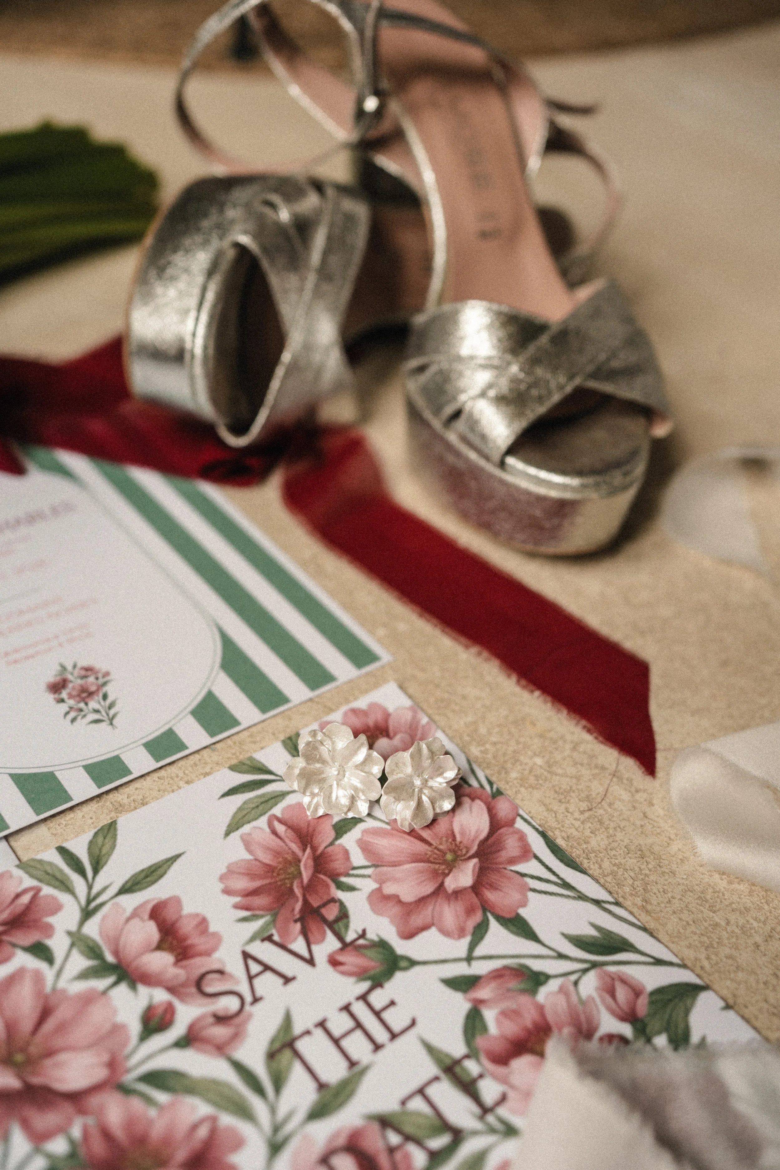 Des chaussures à talons en cuir argenté avec des lacets sur une surface beige, accompagnées de cartes de vœux avec des motifs floraux roses et verts, et des fleurs en plastique blanc.