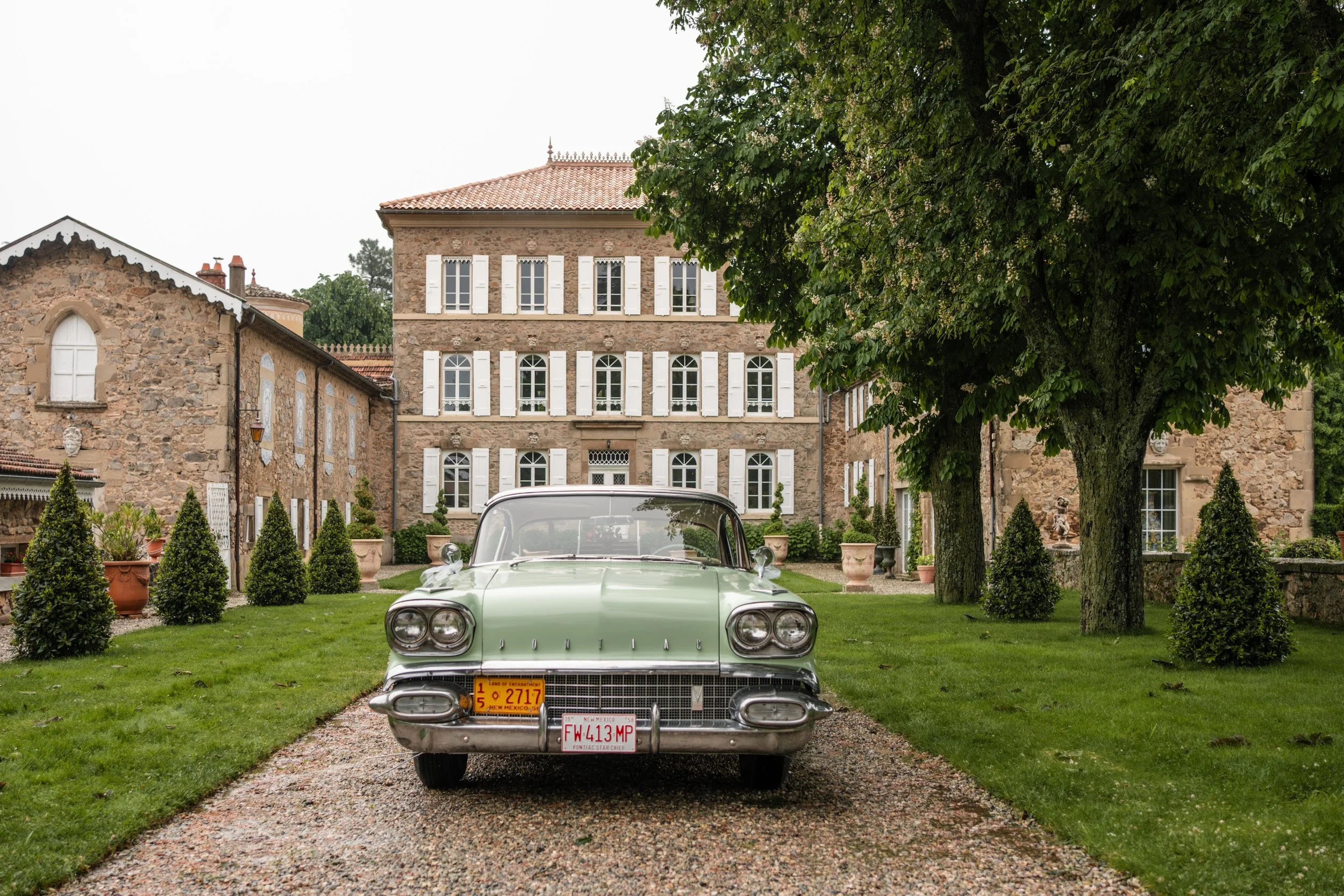 Voiture vintage verte garée sur un chemin en gravier devant une maison en pierre avec volets blancs, entourée d'arbres et de plantes en pot.