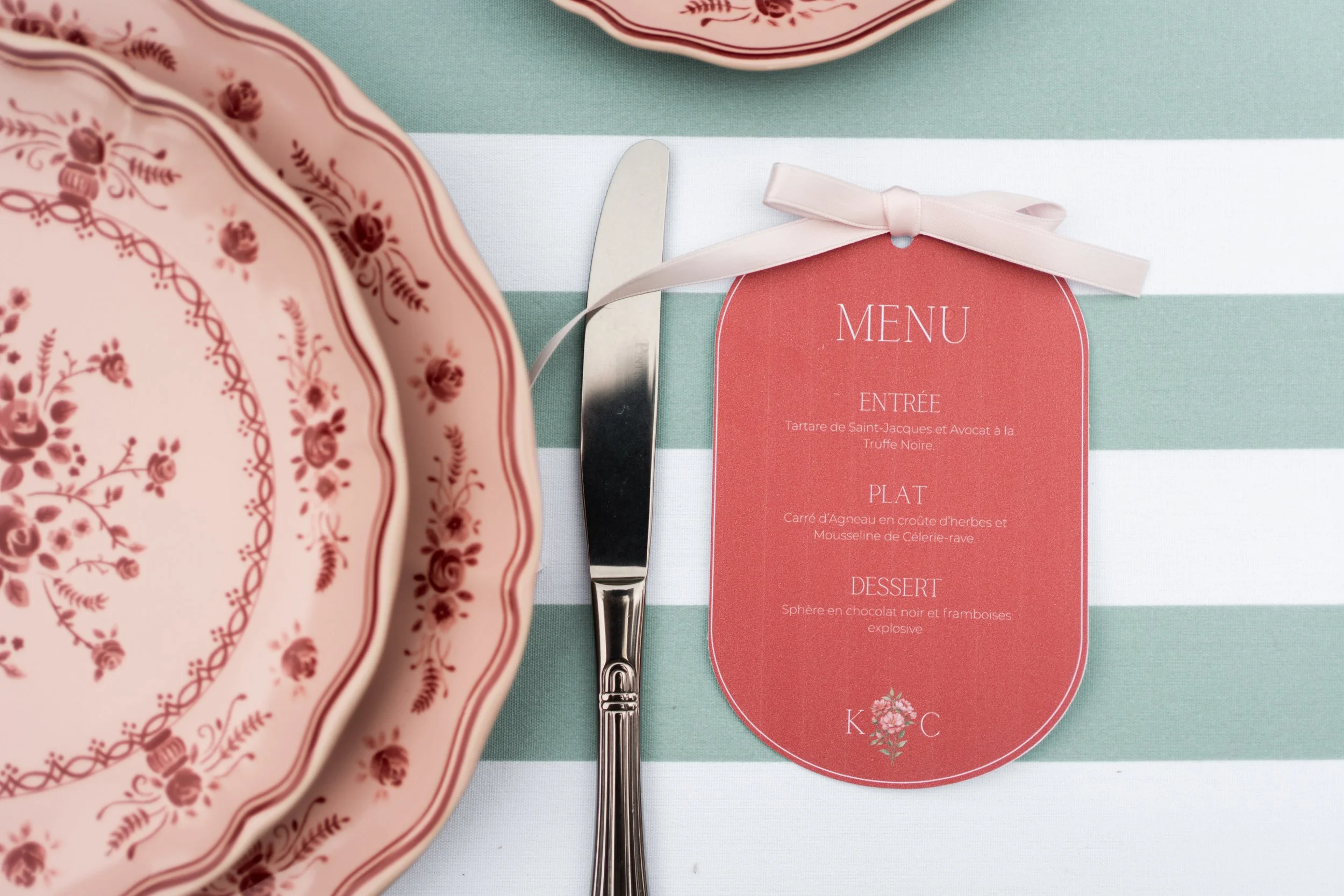 Une assiette en porcelaine rose avec un motif floral en bordure, une cuillère en métal et un menu rouge avec une bande blanche, placé sur une nappe à rayures vertes et blanches.