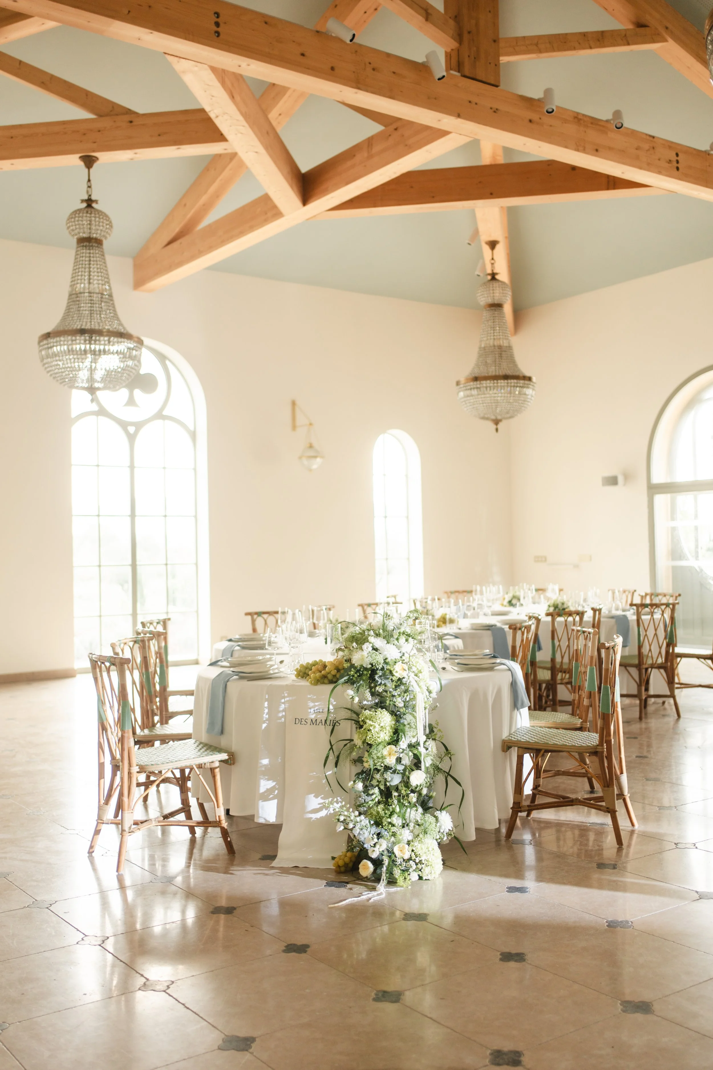 Salle de réception décorée pour un événement, avec une longue table dressée et une fontaine centrale avec des fleurs et des grappes de raisin. Murs blancs, grandes fenêtres, plafonds en bois avec poutres apparentes, et lustres en cristal.