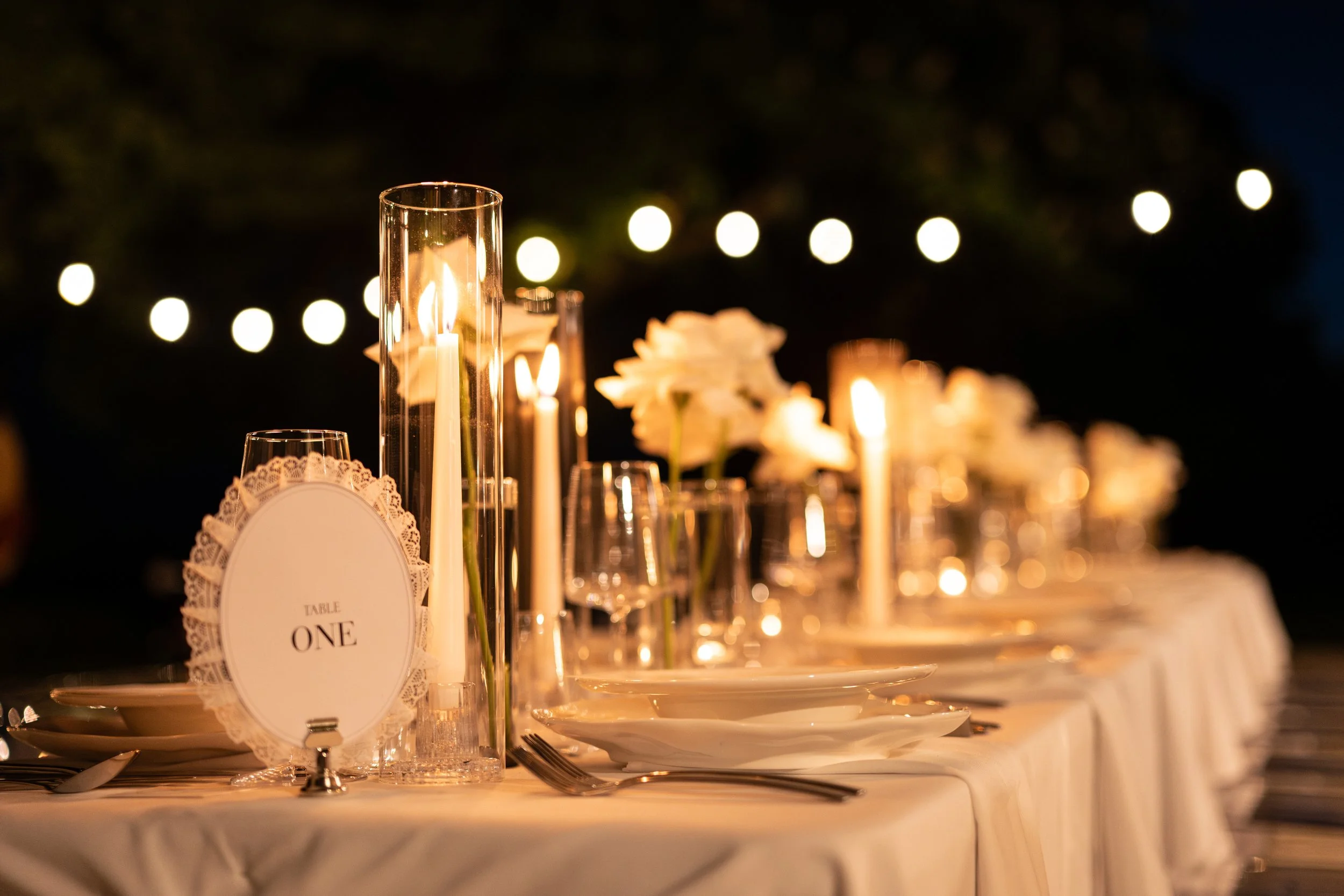 Une table élégante est décorée avec des bougies allumées dans des photophores en verre, entourée de fleurs blanches et de vaisselle raffinée, avec des guirlandes lumineuses au fond créant une ambiance chaleureuse et festive.