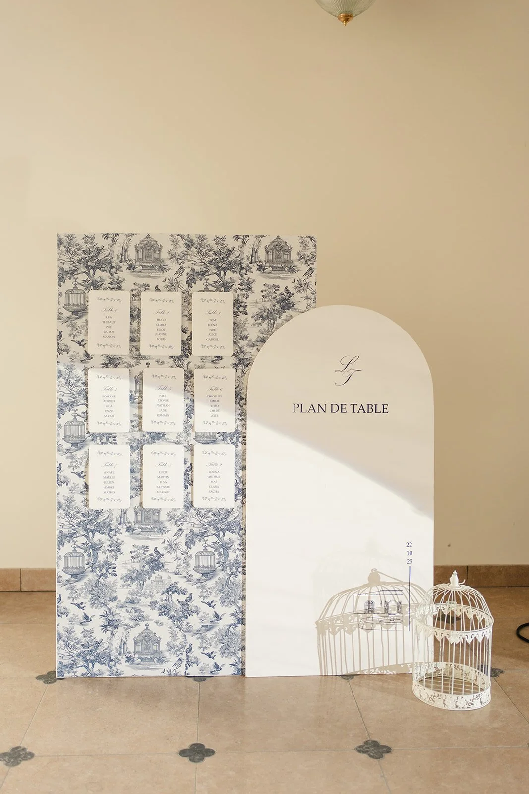 Plan de table pour un événement, avec un fond en bleu et blanc de style toile de Jouy, affichant les noms et numéros de table, à côté d'une grande affiche blanche avec l'inscription 'Plan de table' et une initiale 'G', accompagné de deux cages blanch