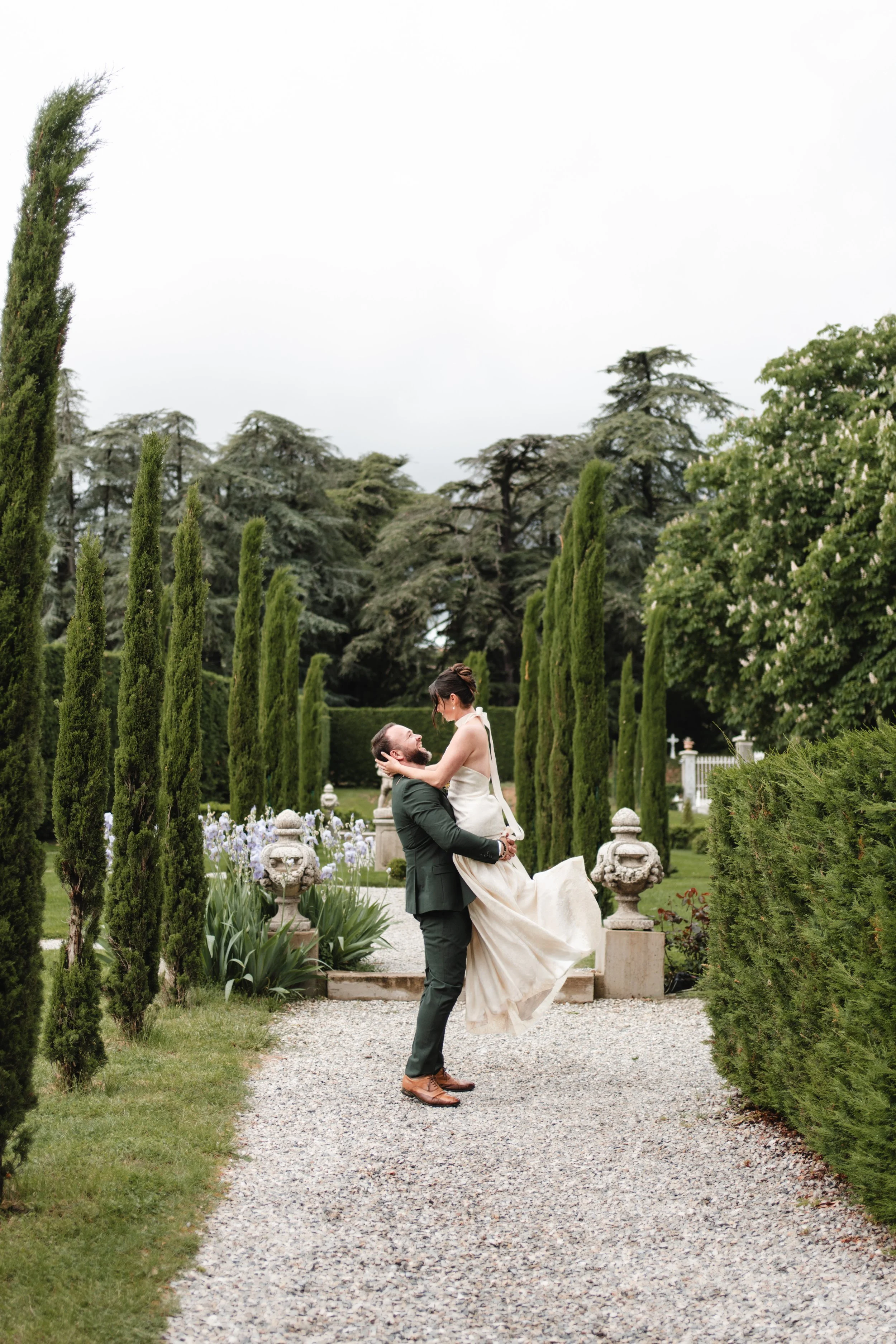 Un homme en costume tient une femme en robe de mariée dans un jardin verdoyant, entouré d'arbres et de sculptures.