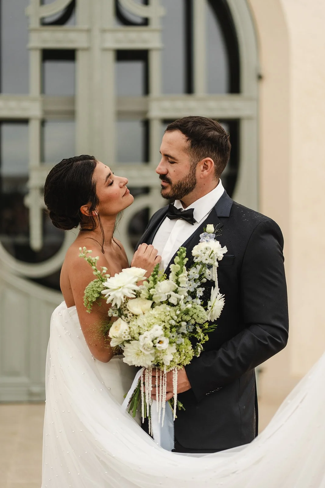 Un couple en tenue de mariage regarde tendrement l'un l'autre, la femme tient un bouquet de fleurs blanches, dans une salle élégante.