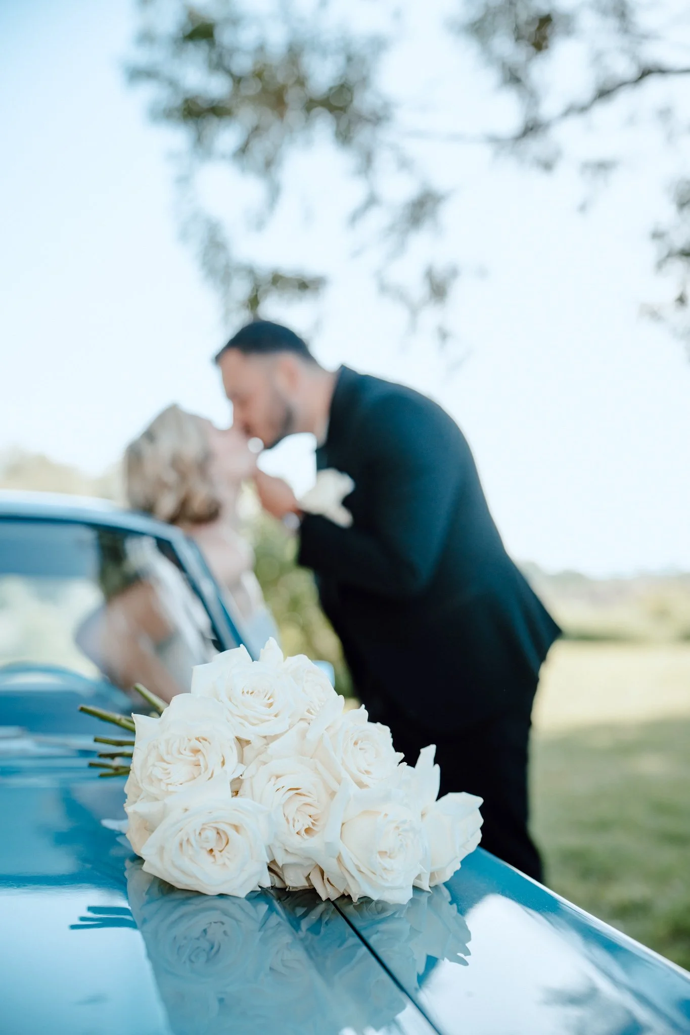 Un couple échange un baiser sur le capot de leur voiture décorée avec un bouquet de roses blanches, lors d'une cérémonie de mariage en plein air.
