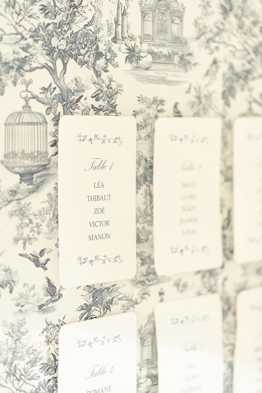 Liste de noms de personnes sur un marqueur de table, avec un fond décoratif en noir et blanc représentant un paysage avec des arbres, des oiseaux et des bâtiments classiques.