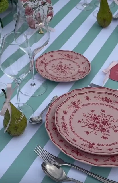 Table dressée pour un repas avec des assiettes en porcelaine rouge et blanche, des verres à vin, des cuillères, des fourchettes, des poires décoratives, et un chemin de table à rayures vert et blanc.