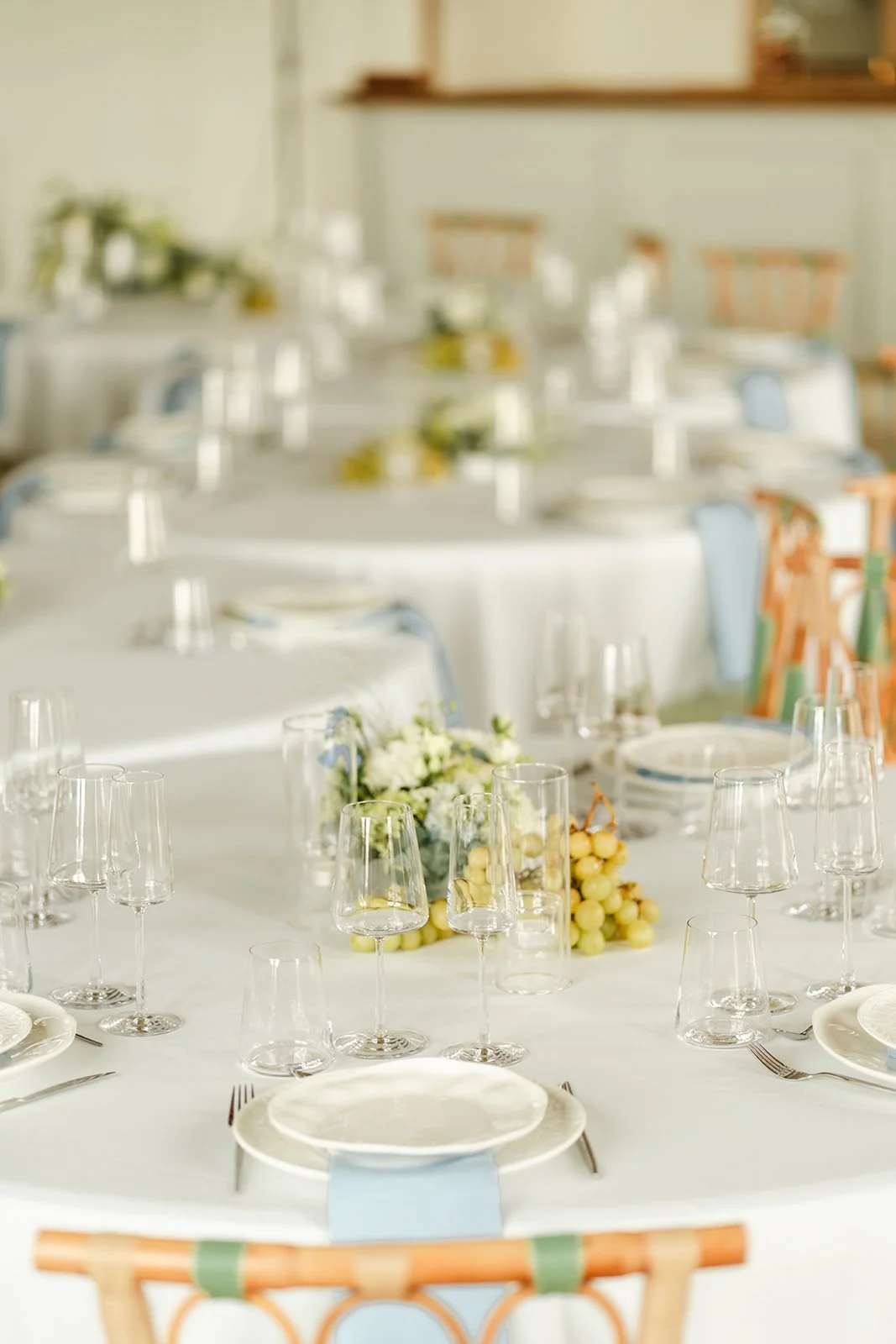 Table de fête élégamment dressée avec verres à vin, assiettes blanches, décor floral et raisins, dans une salle lumineuse.