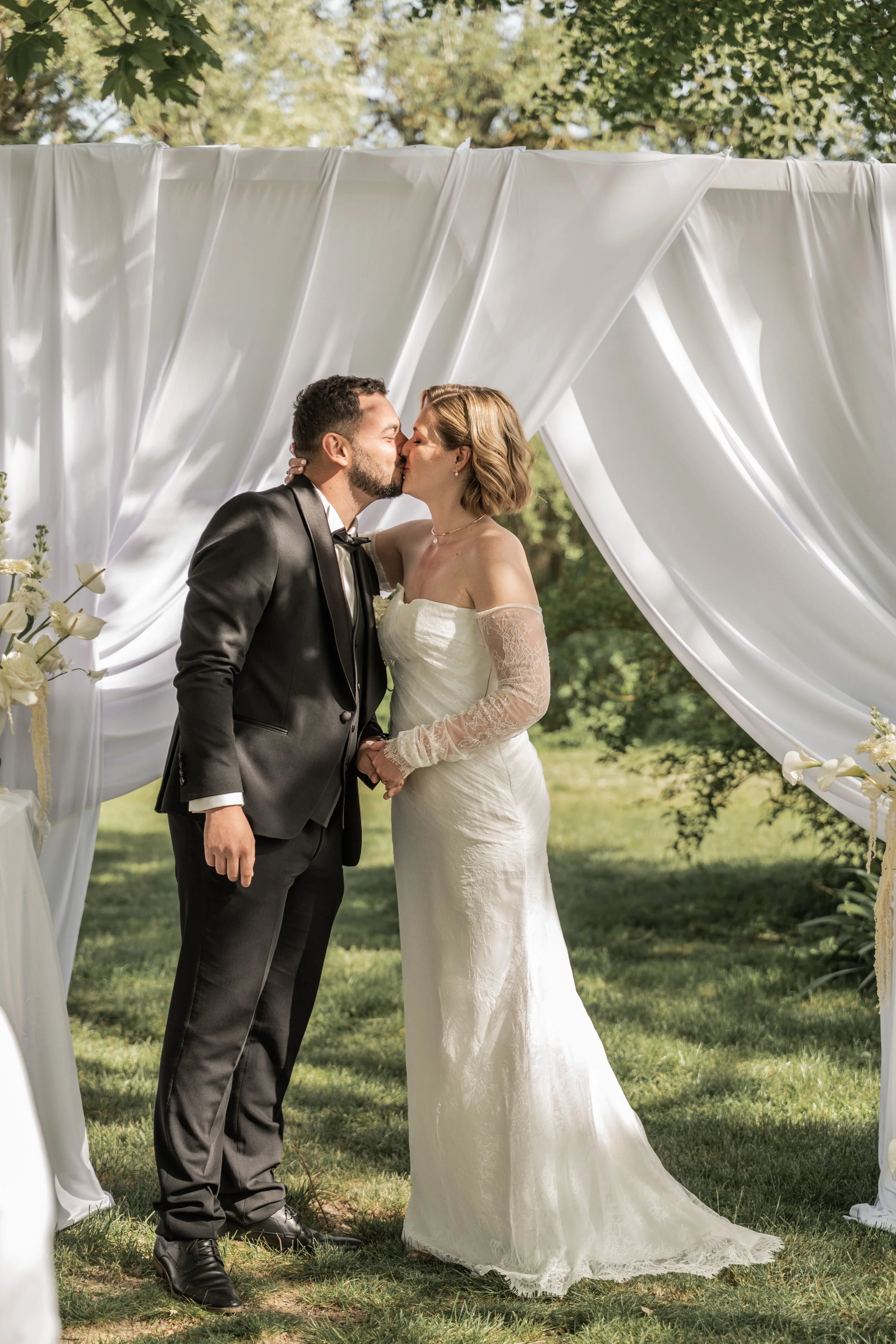 Un couple de mariés s'embrassant lors de leur cérémonie en plein air, sous un décor de tissu blanc drapé, avec des arbres en arrière-plan.