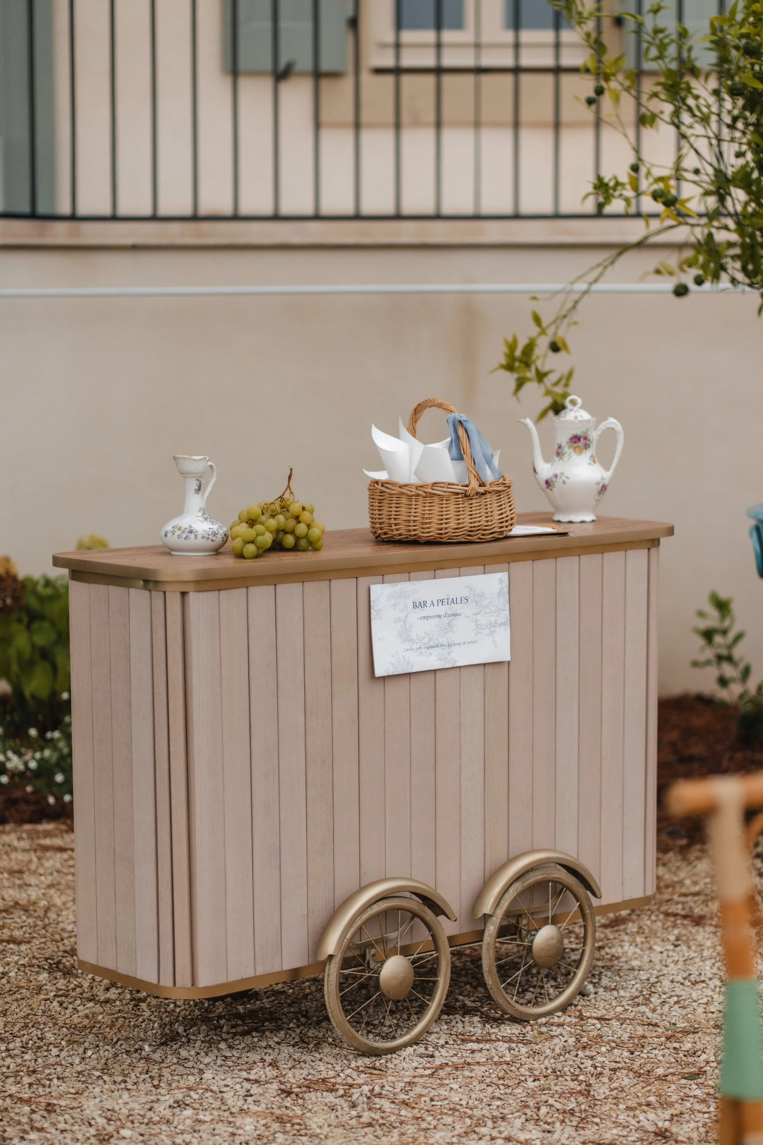 Un chariot en bois ressemblant à une boutique de vente de fleurs ou de boissons, décoré de vases, raisin et un panier avec des papiers ou serviettes, en extérieur sur un sol en gravier.