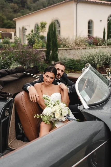 Un couple de mariés assis dans une voiture décapotable devant une maison avec jardin.