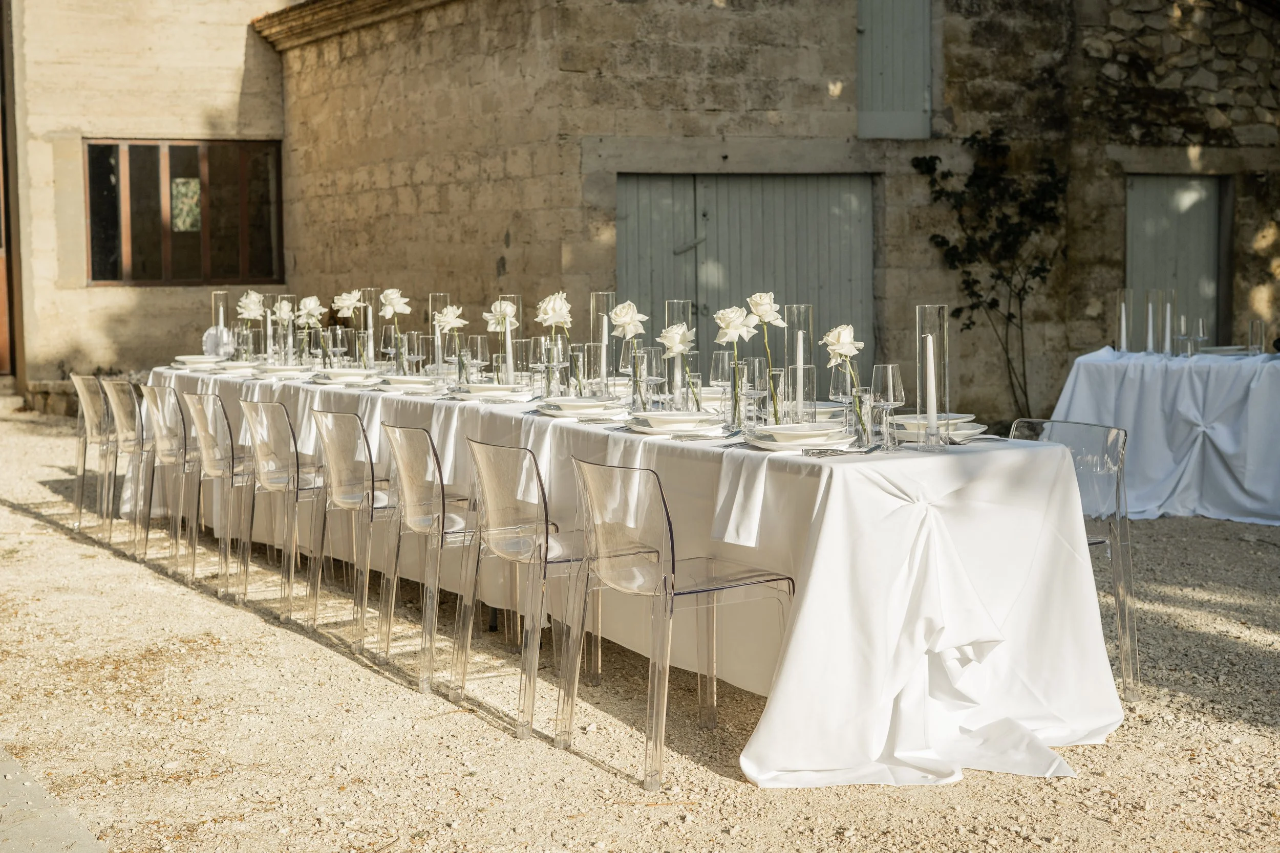 Une longue table dressée avec une nappe blanche, ornée de vases avec des fleurs blanches, dans une cour en pierre avec des fenêtres en bois, pour un événement ou un repas en plein air.