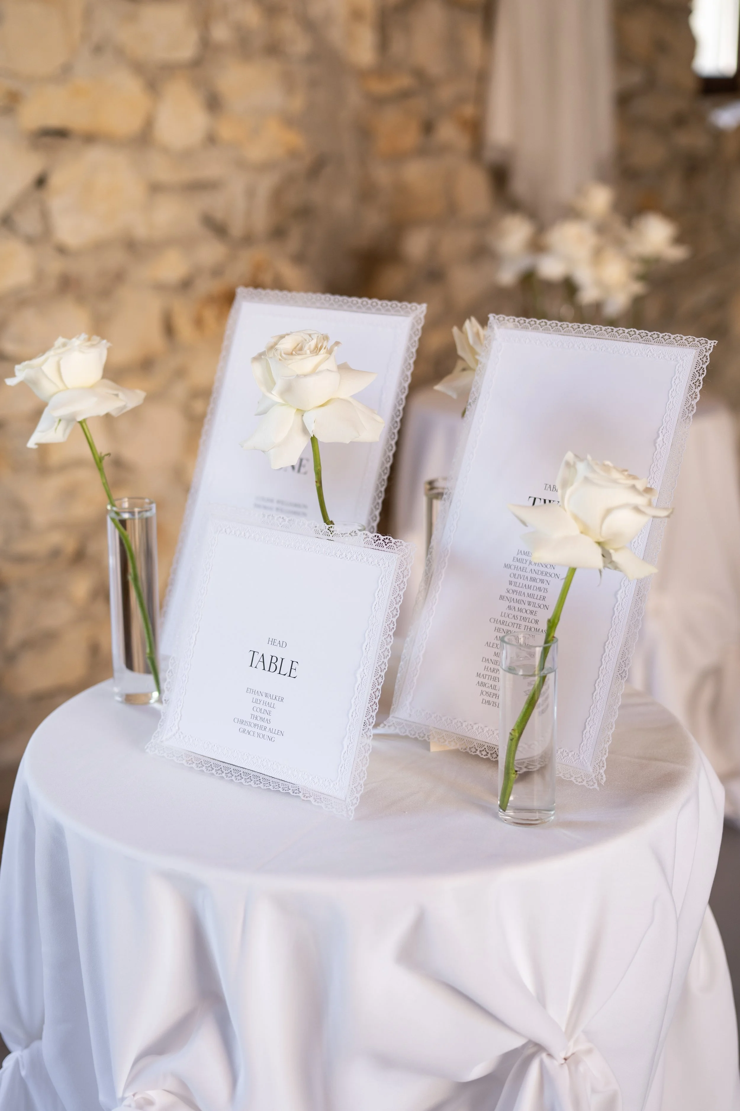Tables avec des menus de mariage ornés de roses blanches dans des vases en verre, placés sur une table ronde recouverte d'une nappe blanche, avec un mur en pierre en arrière-plan.