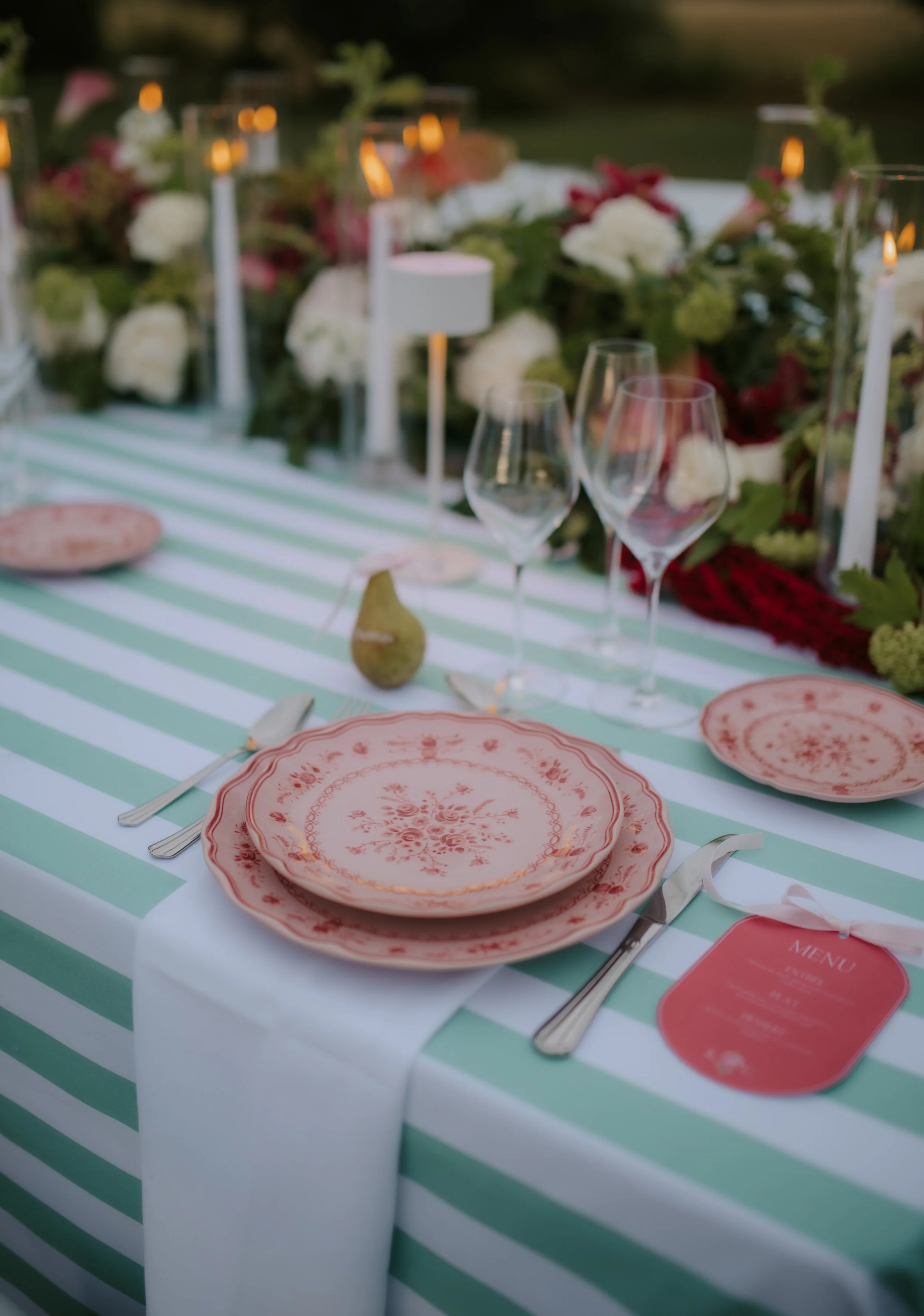 Table de fête décorée avec des assiettes en porcelaine rose, des verres à vin, des couverts en métal, une figue, et une nappe à rayures vertes et blanches, entourée de fleurs et de bougies électriques dans un jardin en soirée.