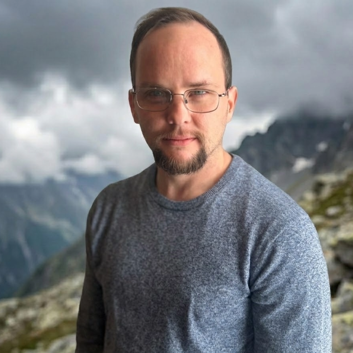 A man with glasses and a beard standing outdoors in front of mountains and cloudy sky.